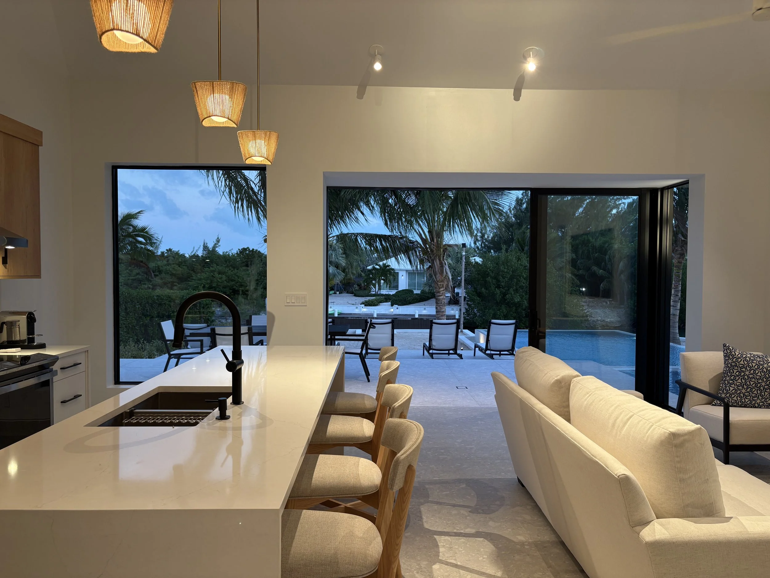 Living room with open kitchen, island with bar stools, beige sofa, and sliding glass door leading to outdoor pool area with palm trees, chairs, and patio.