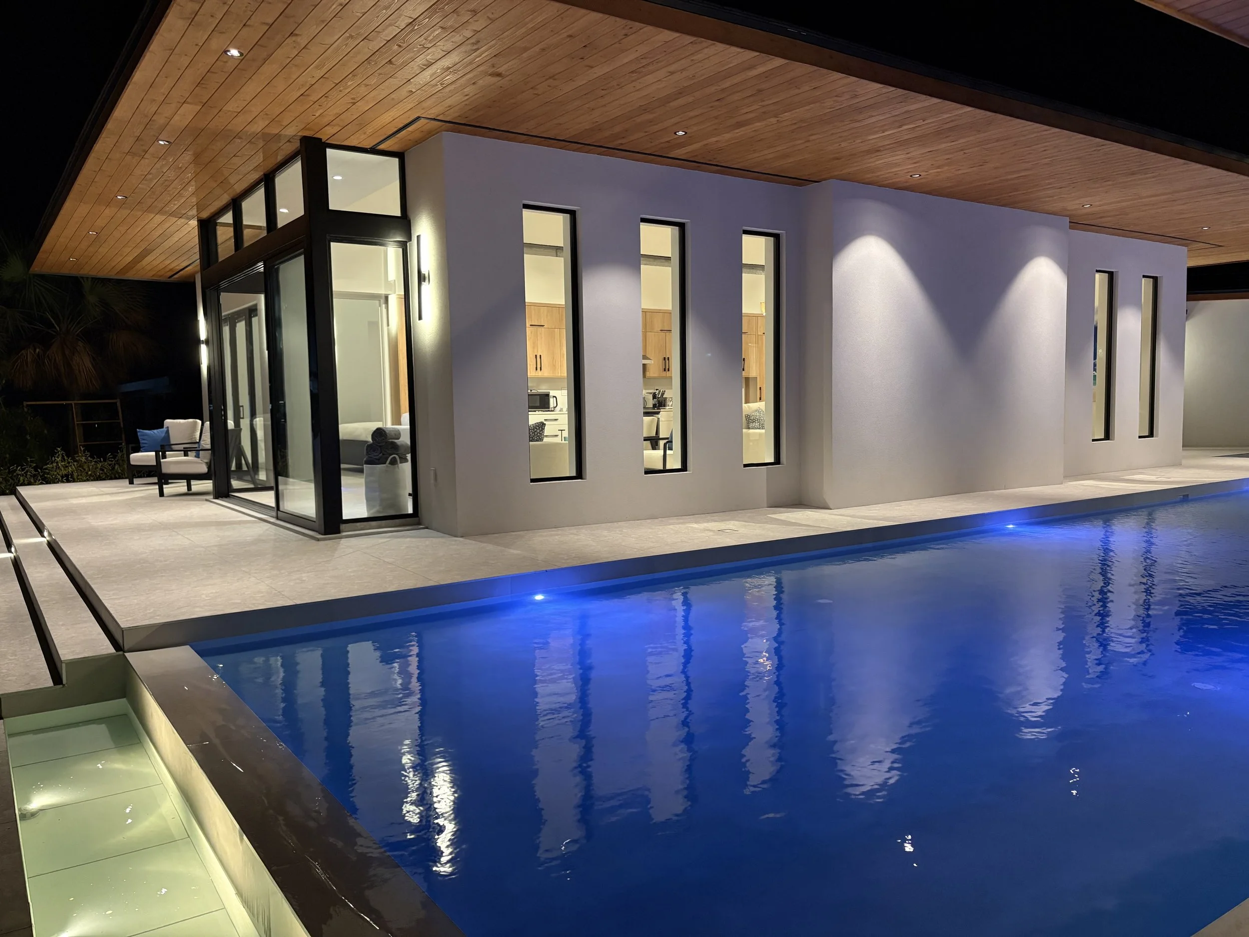 Modern house with large windows and a swimming pool at night, illuminated by exterior lighting, with a wooden ceiling over the patio.