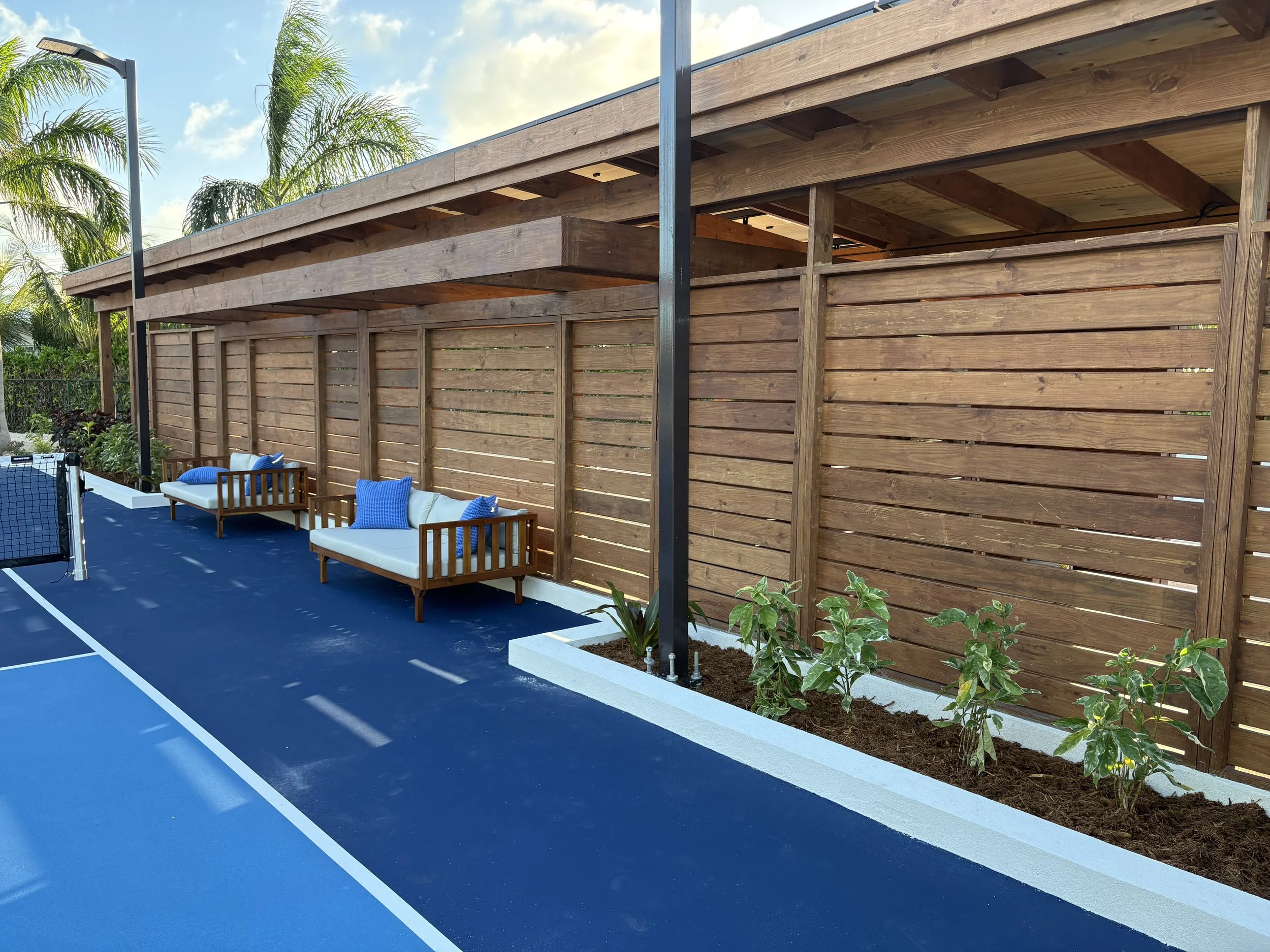 A backyard with a blue tennis court, two wooden benches with white cushions and blue pillows, and a tall wooden privacy fence with plants in front.