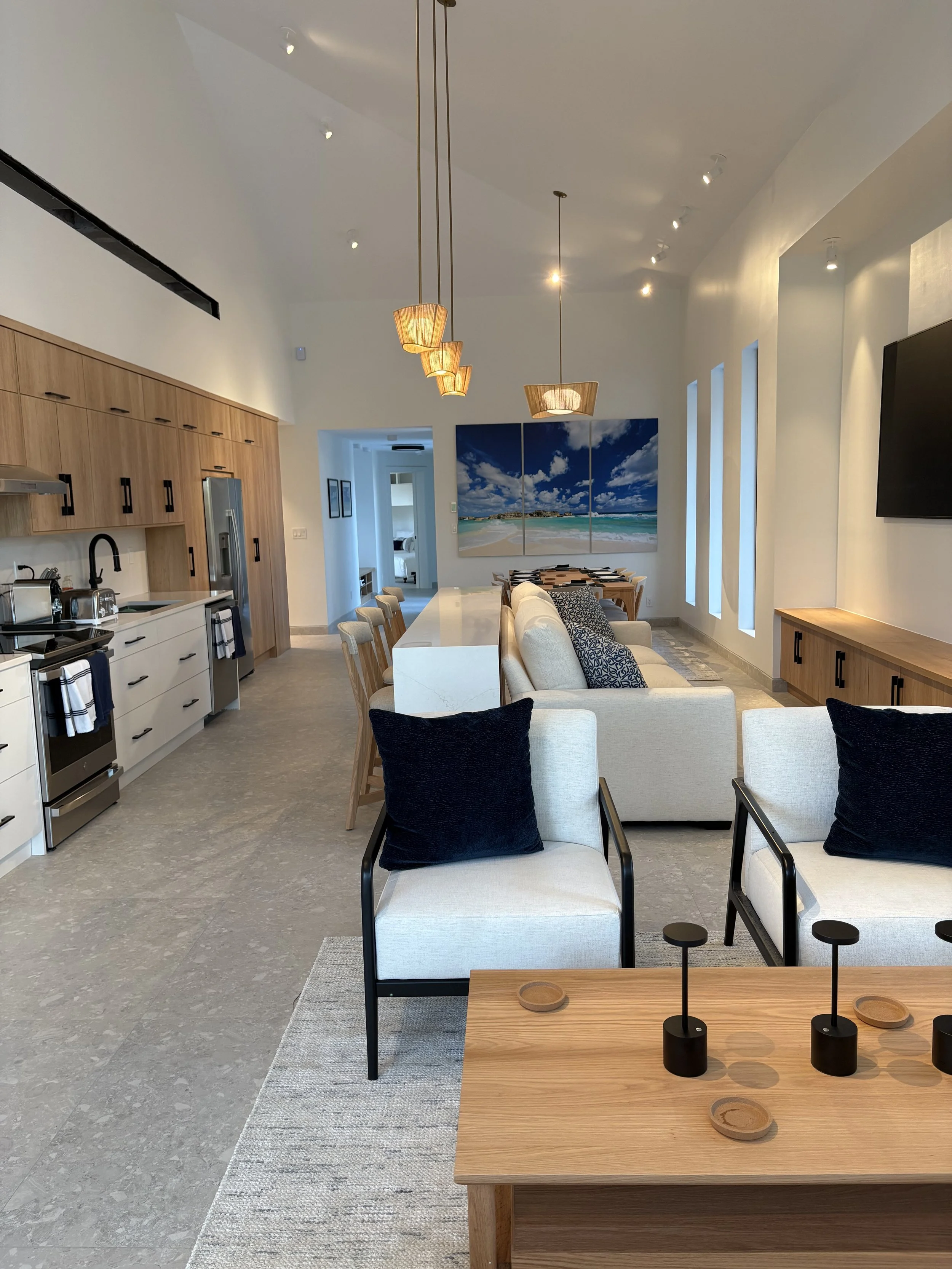 Modern open-concept living room with a kitchen area, beige sofa with black pillows, dining area, and wall art depicting a beach scene.