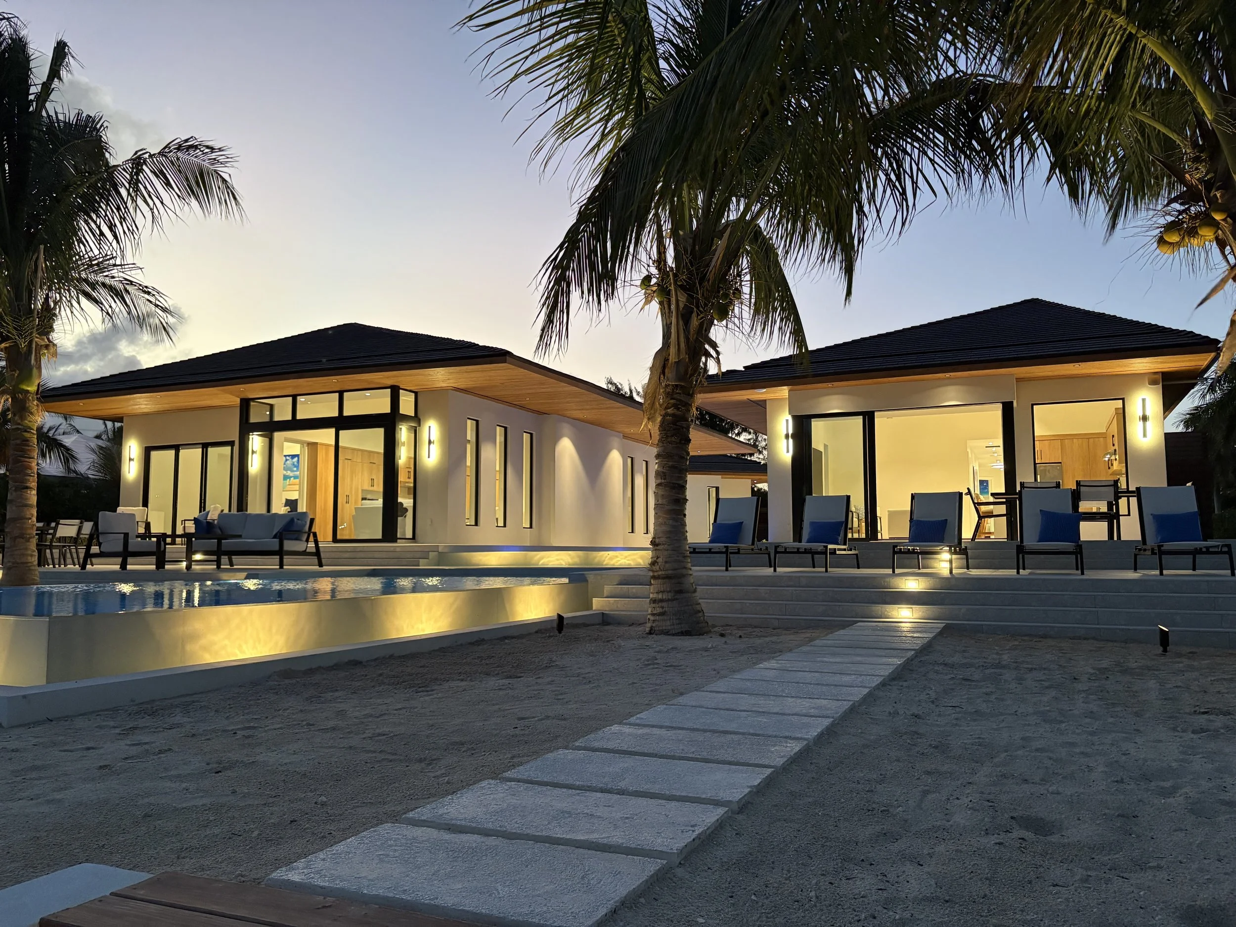 Modern house with large glass windows and sliding doors, illuminated outdoor lighting, a small swimming pool, lounge chairs, and palm trees in the evening.