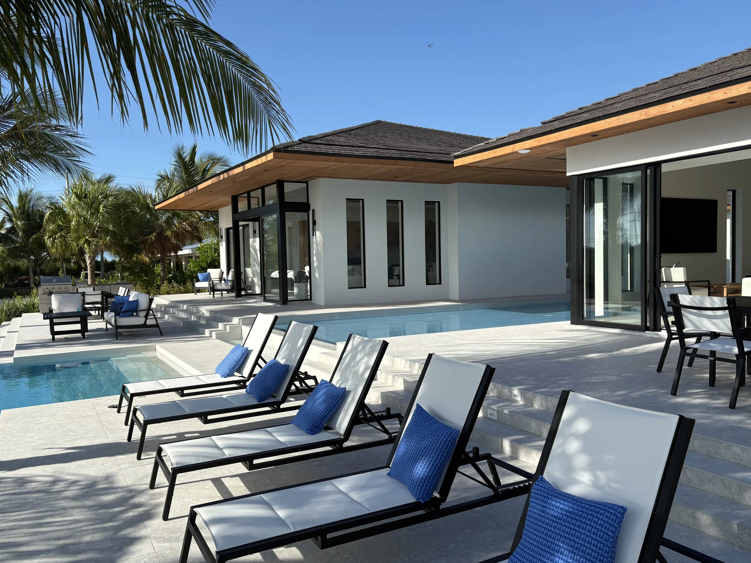 Luxury modern house with private pool, outdoor lounge chairs with blue cushions, tropical palm trees, and clear blue sky