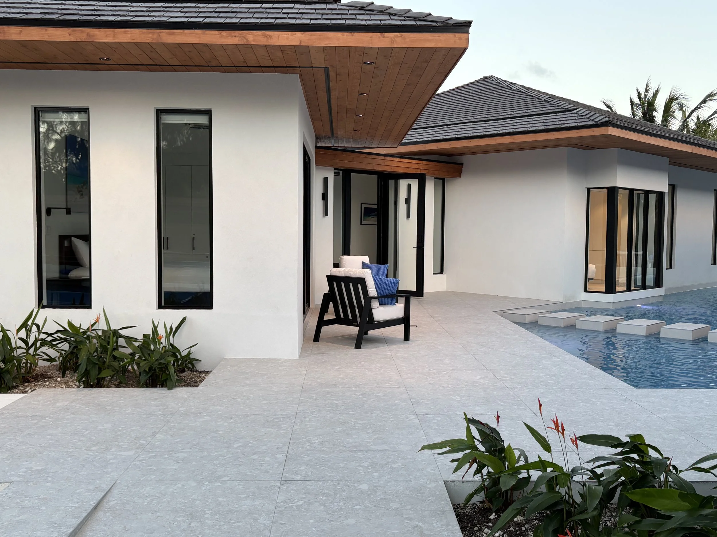 Modern house with white exterior walls, large glass doors and windows, wooden accents on the ceiling and roof, outdoor seating with cushions, and a swimming pool with stepping stones.