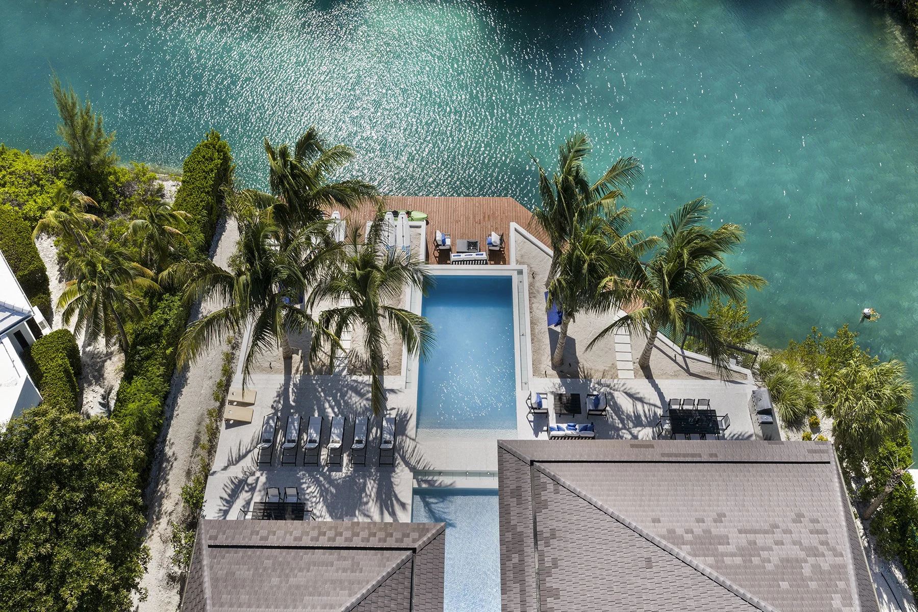 Aerial view of a tropical waterfront property with a swimming pool, palm trees, outdoor furniture, and a deck area overlooking a calm body of water.