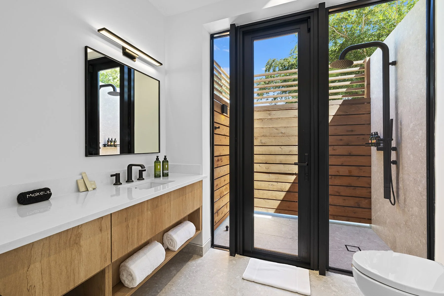 Modern bathroom with white walls, wooden vanity with rolled towels, black-framed mirror and door, outdoor shower area with wooden privacy wall, and clear sky visible through the glass door.