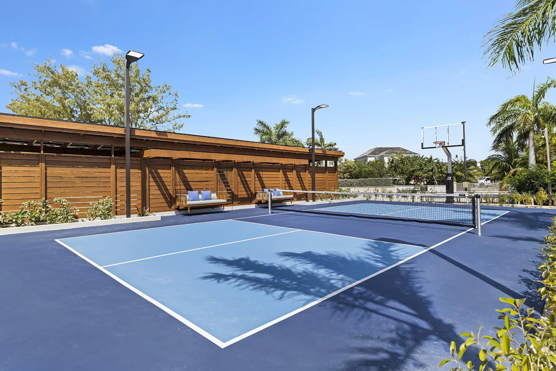 An outdoor tennis court with a blue playing surface, a net, and a basketball hoop, surrounded by palm trees and a wooden fence, under a clear blue sky.
