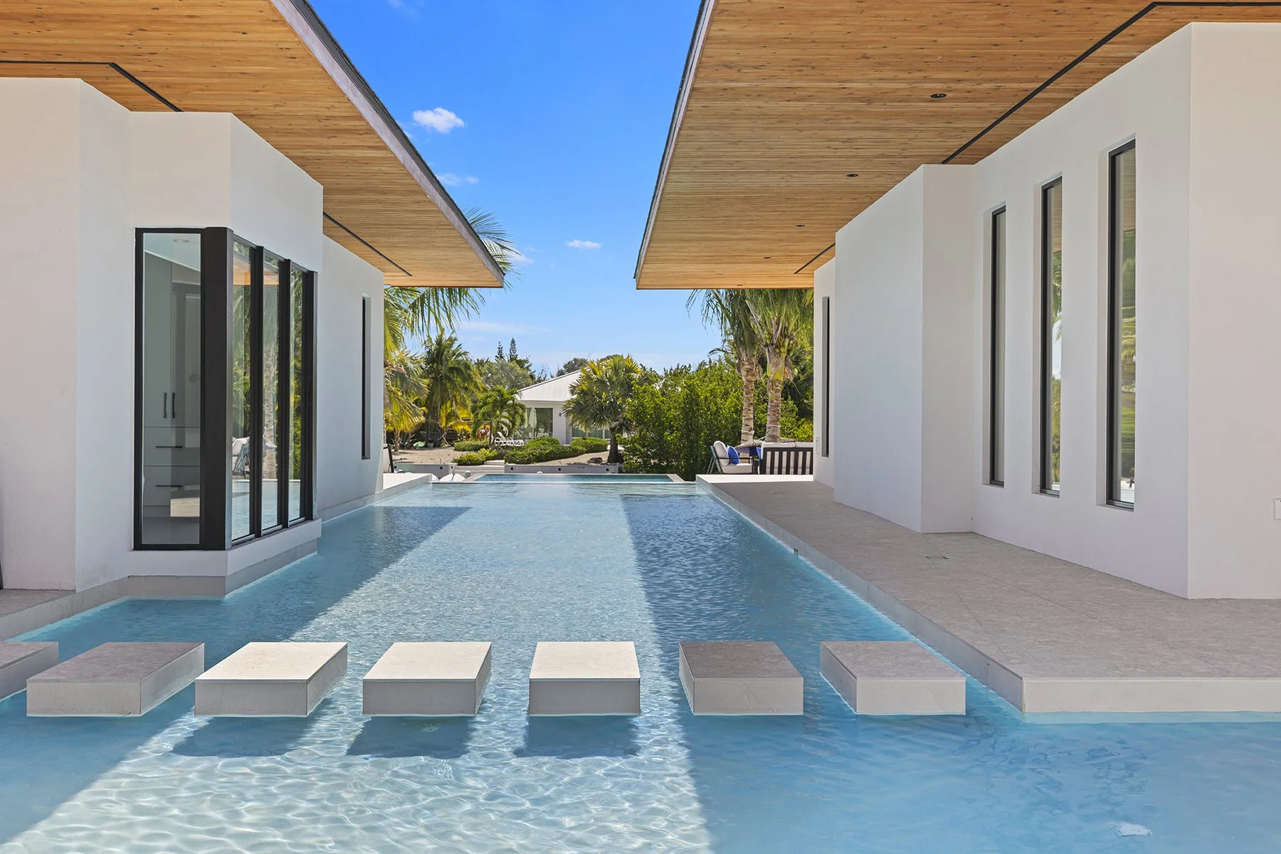 Modern house with a swimming pool and stepping stones, surrounded by palm trees and lush greenery, under a clear blue sky.