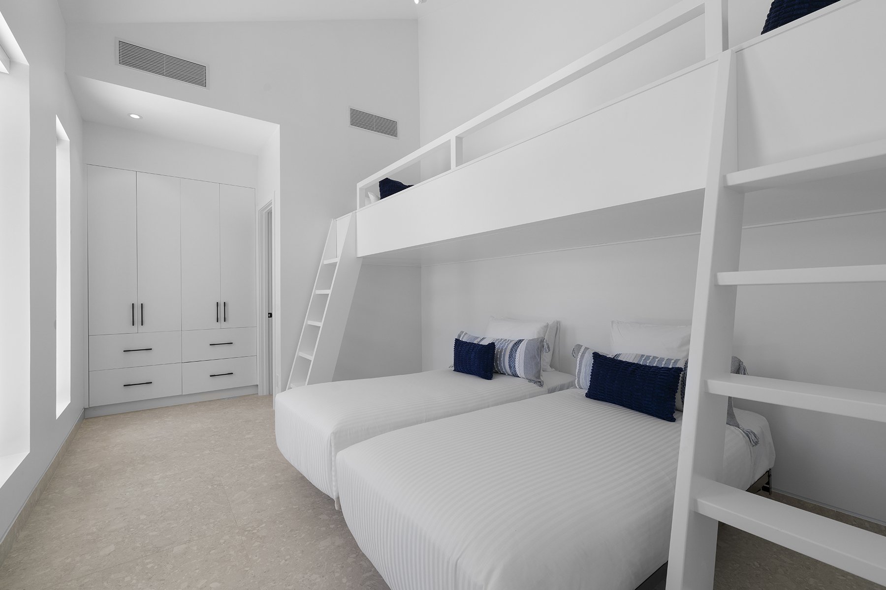 Room with two beds, white linens, decorative pillows, and a loft with a ladder, all in a modern white interior.