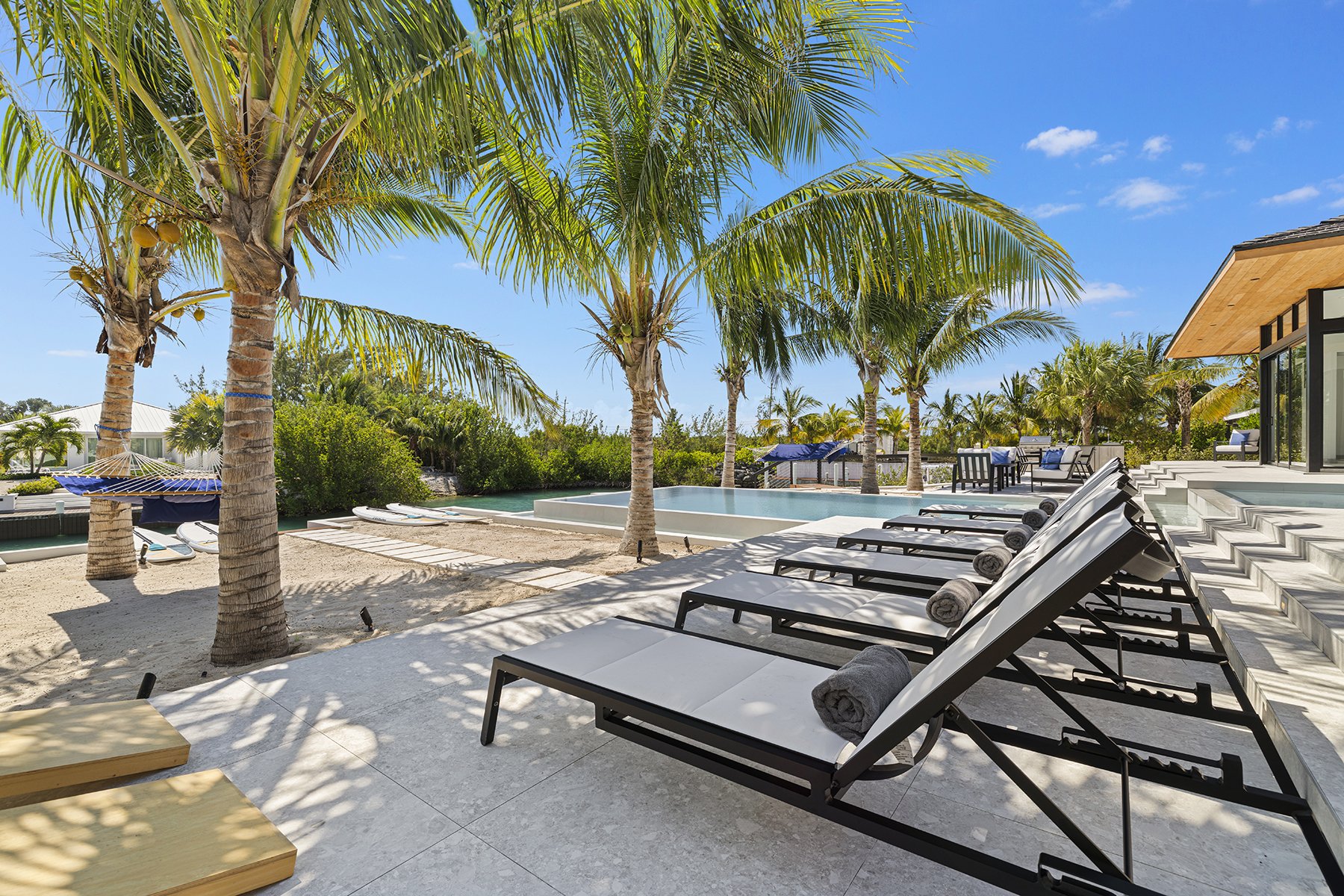 Luxurious outdoor pool area with black lounge chairs, rolled towels, palm trees, and a sandy beach area under a bright blue sky.