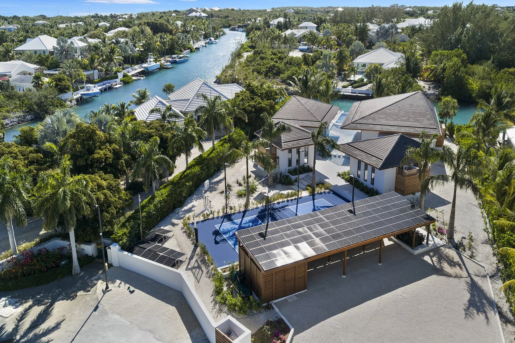A luxury waterfront villa with solar panels on the roof, surrounded by lush palm trees, a driveway, and a covered area with solar panels. In the background, there are houses along waterways with boats docked nearby, lush greenery, and clear blue wate