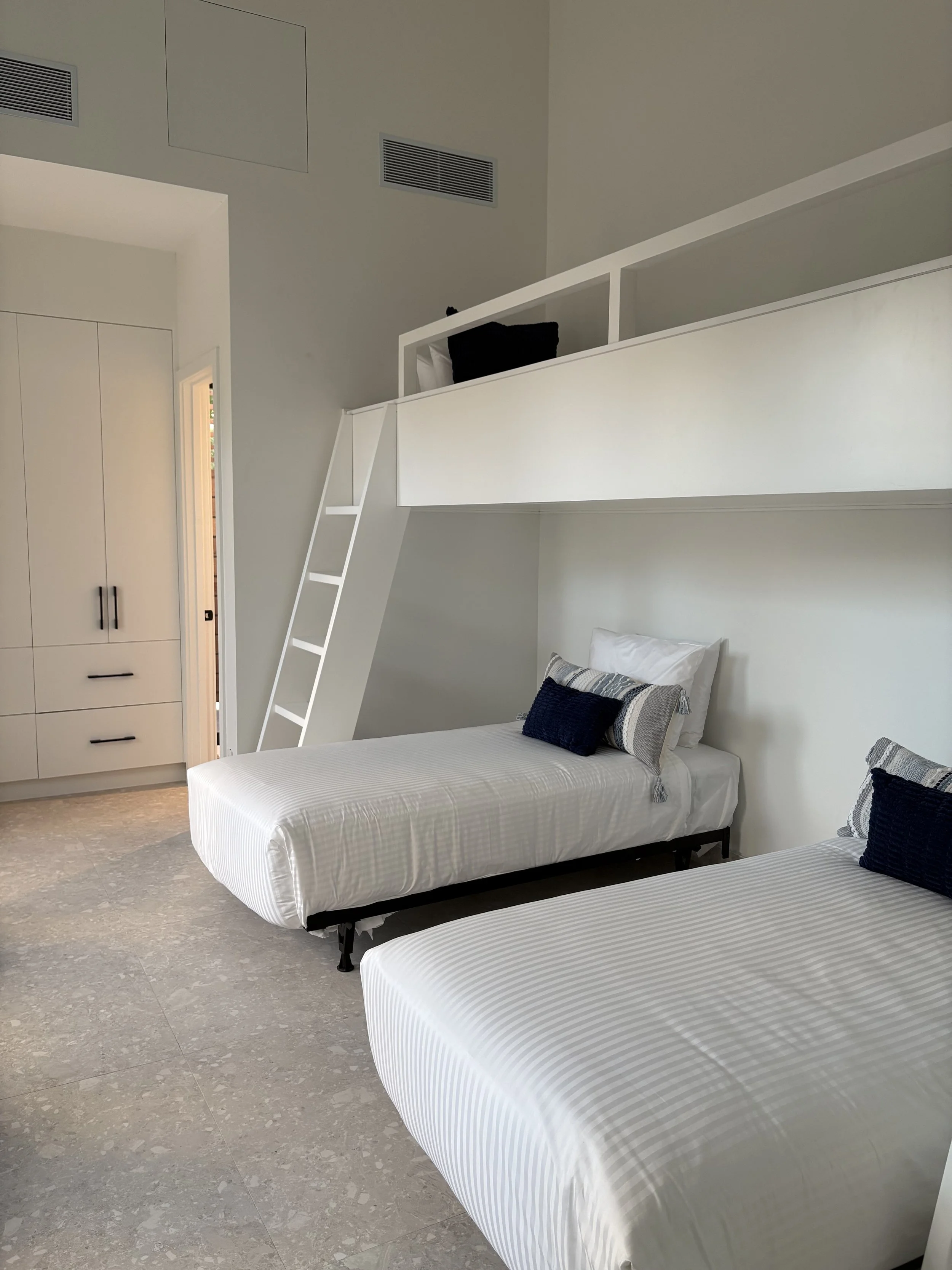 Bedroom with two single beds covered in white striped bedding, each with navy and gray pillows. A loft bed with white railing and a ladder is above the beds. The room has white walls, a built-in wardrobe, and a small door leading to a closet or bathr