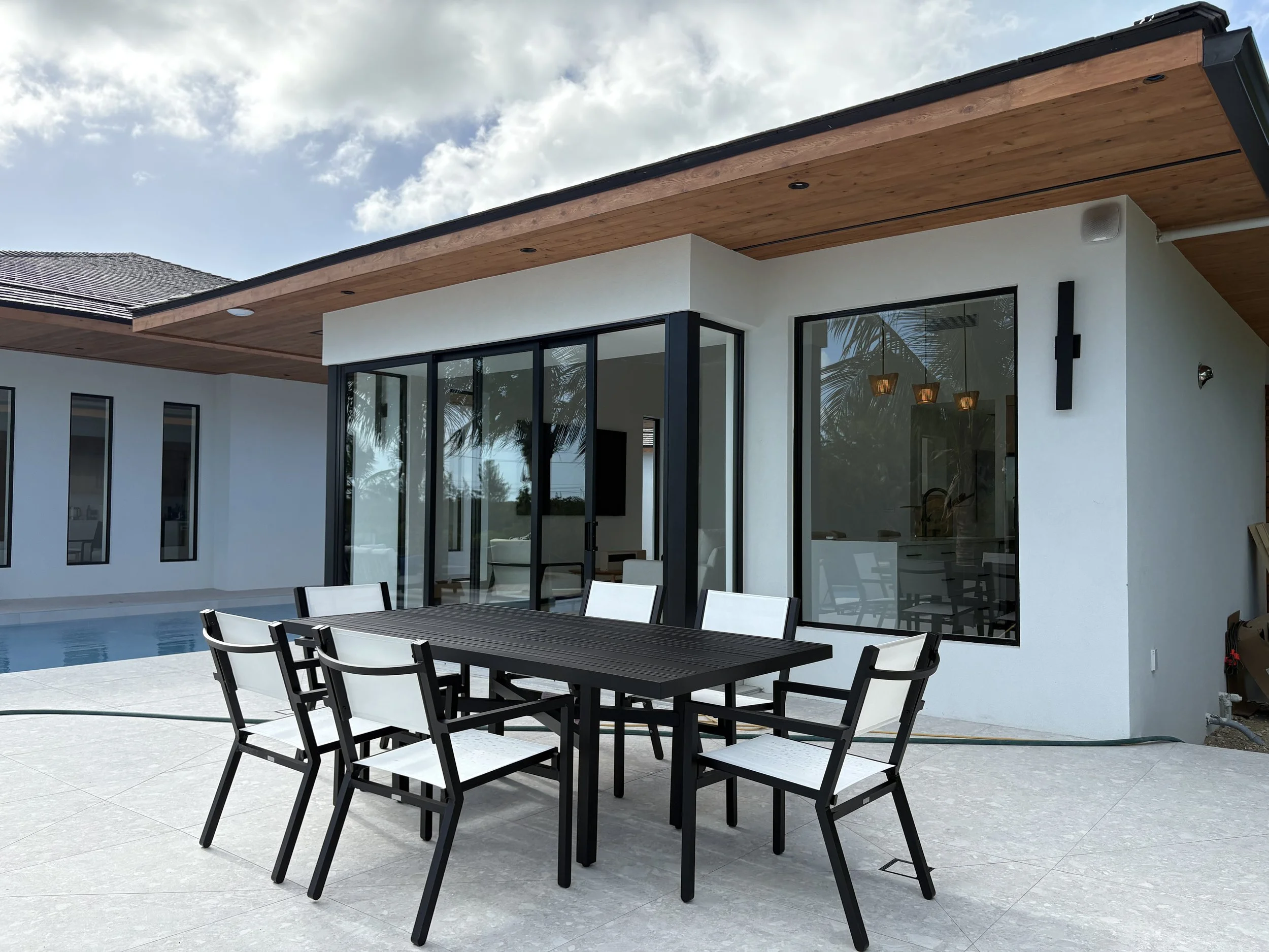 Modern house with large glass sliding doors, outdoor dining table with six chairs, swimming pool, and tropical palm trees reflected in the windows.