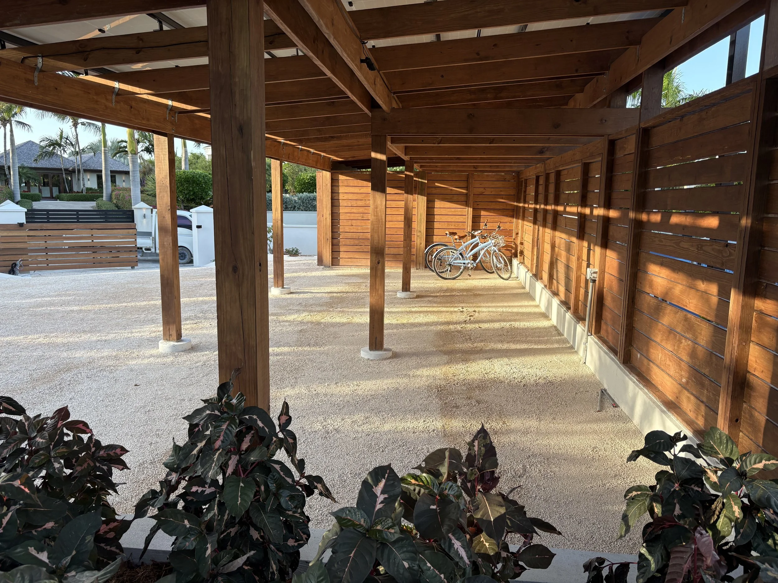 A covered bike parking area with wooden posts and slats, three bicycles parked inside, and shrubbery in the foreground with houses, trees, and a car in the background.