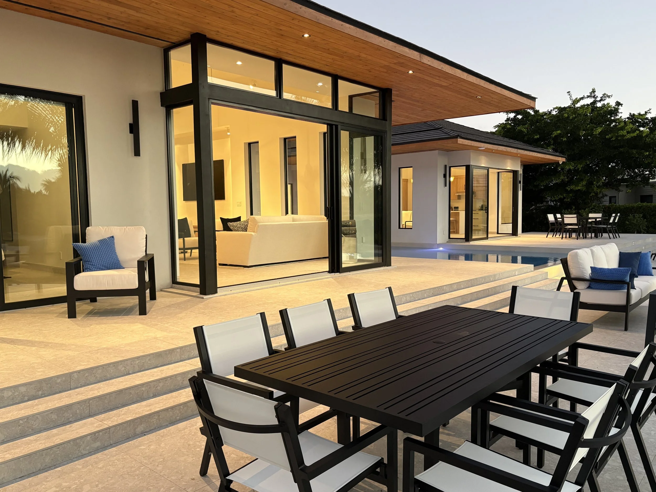 Modern backyard with outdoor dining table and chairs, patio steps, lounge area with sofas and armchairs, all illuminated during dusk, with a pool and trees in the background.
