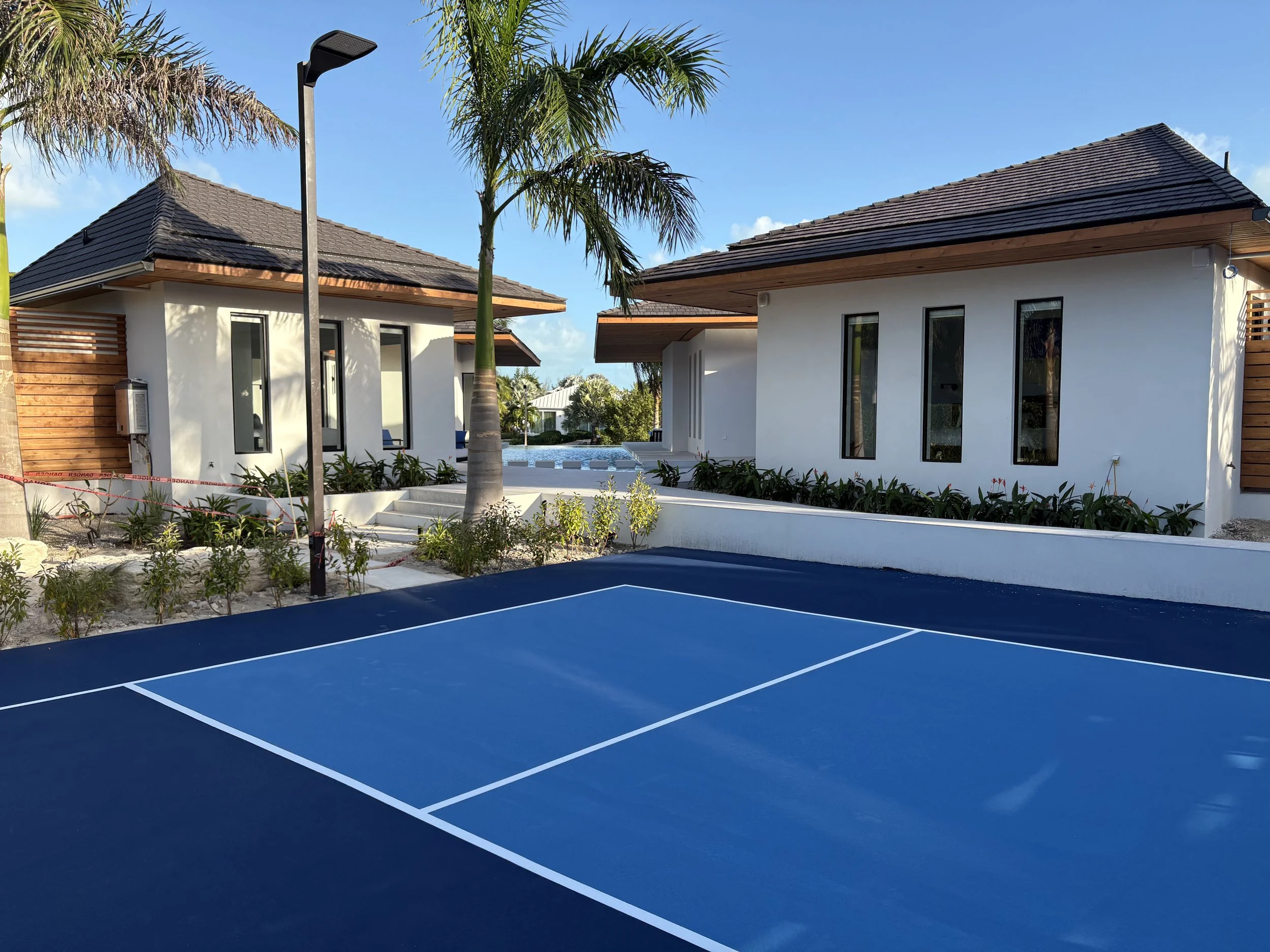 A blue tennis court with white lines in front of two modern white houses with dark roofs, palm trees, and landscaped greenery.