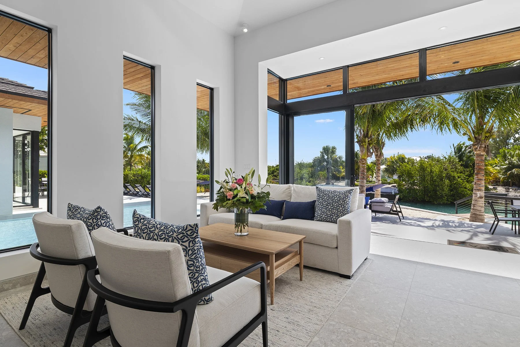 Modern living room with white sofa, patterned pillows, a wooden coffee table with a floral arrangement, and large windows showing outdoor palm trees and a pool area.