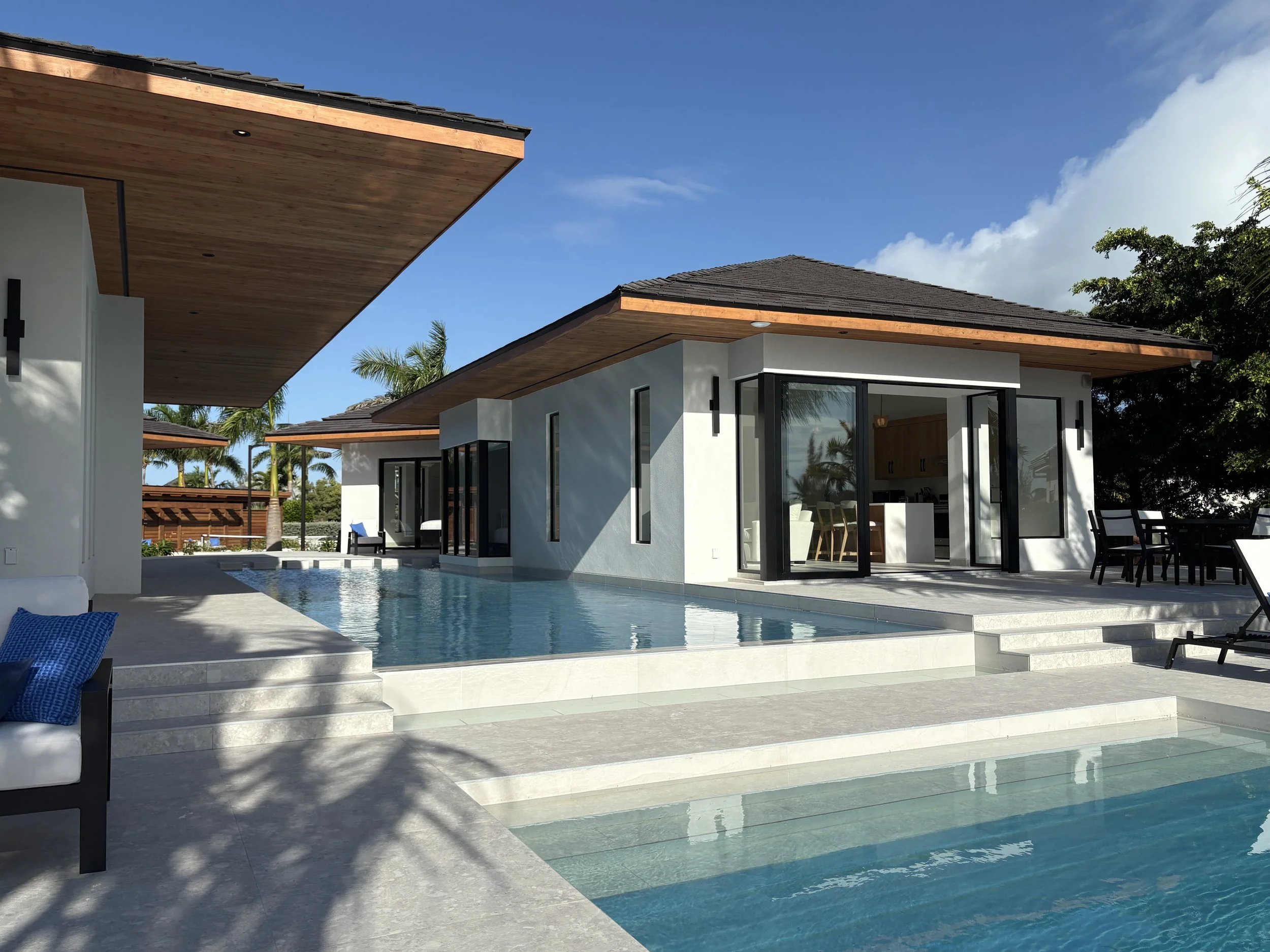 Modern house with a pool in a tropical setting under a clear blue sky, with palm trees in the background and outdoor seating.