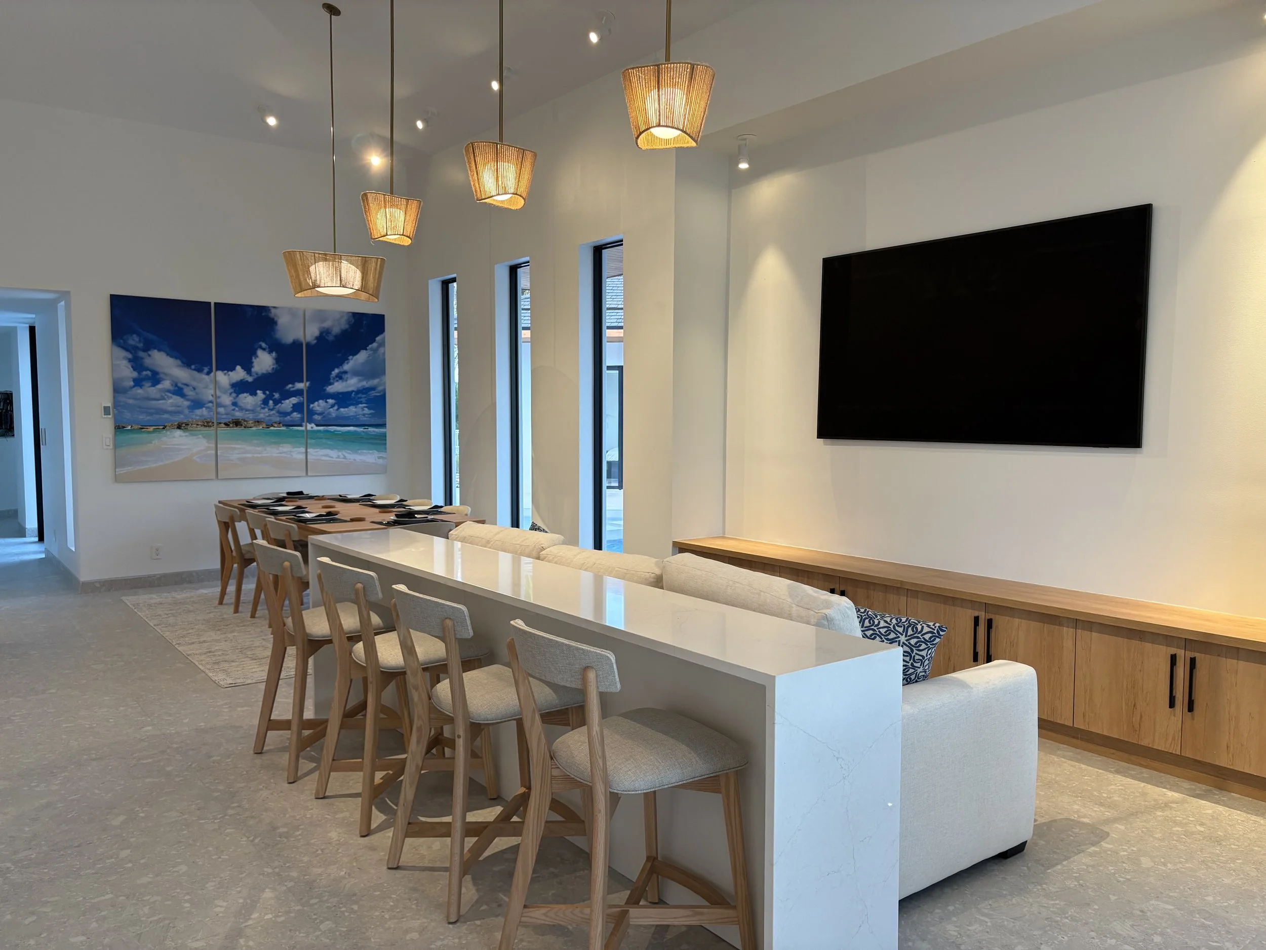 Modern dining and living area with a long white island, beige bar stools, a beige sofa, a large black wall-mounted TV, and a set of three paintings of a beach scene with blue sky and clouds.