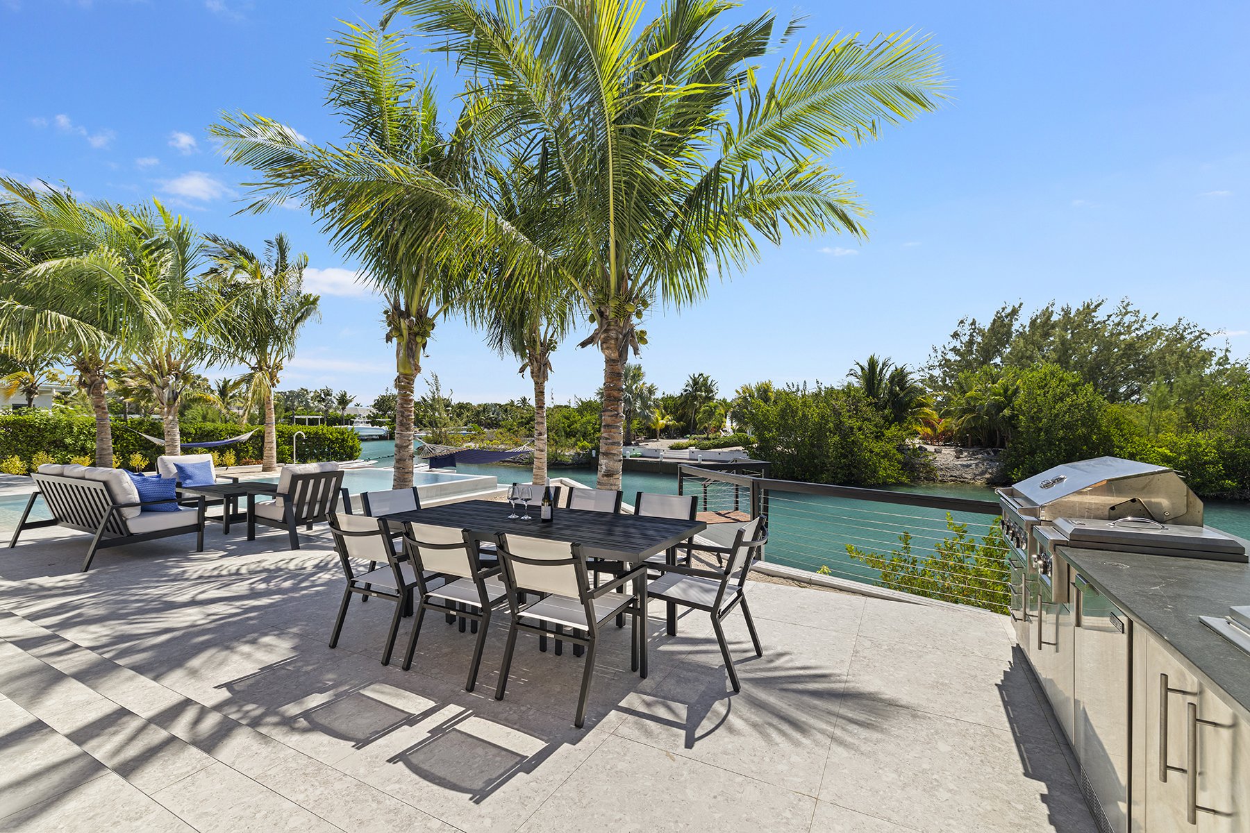 Outdoor patio with a dining table and chairs, a lounge area with cushioned seats, a built-in grill, palm trees, a waterway with a railing, and lush green bushes under a bright blue sky.