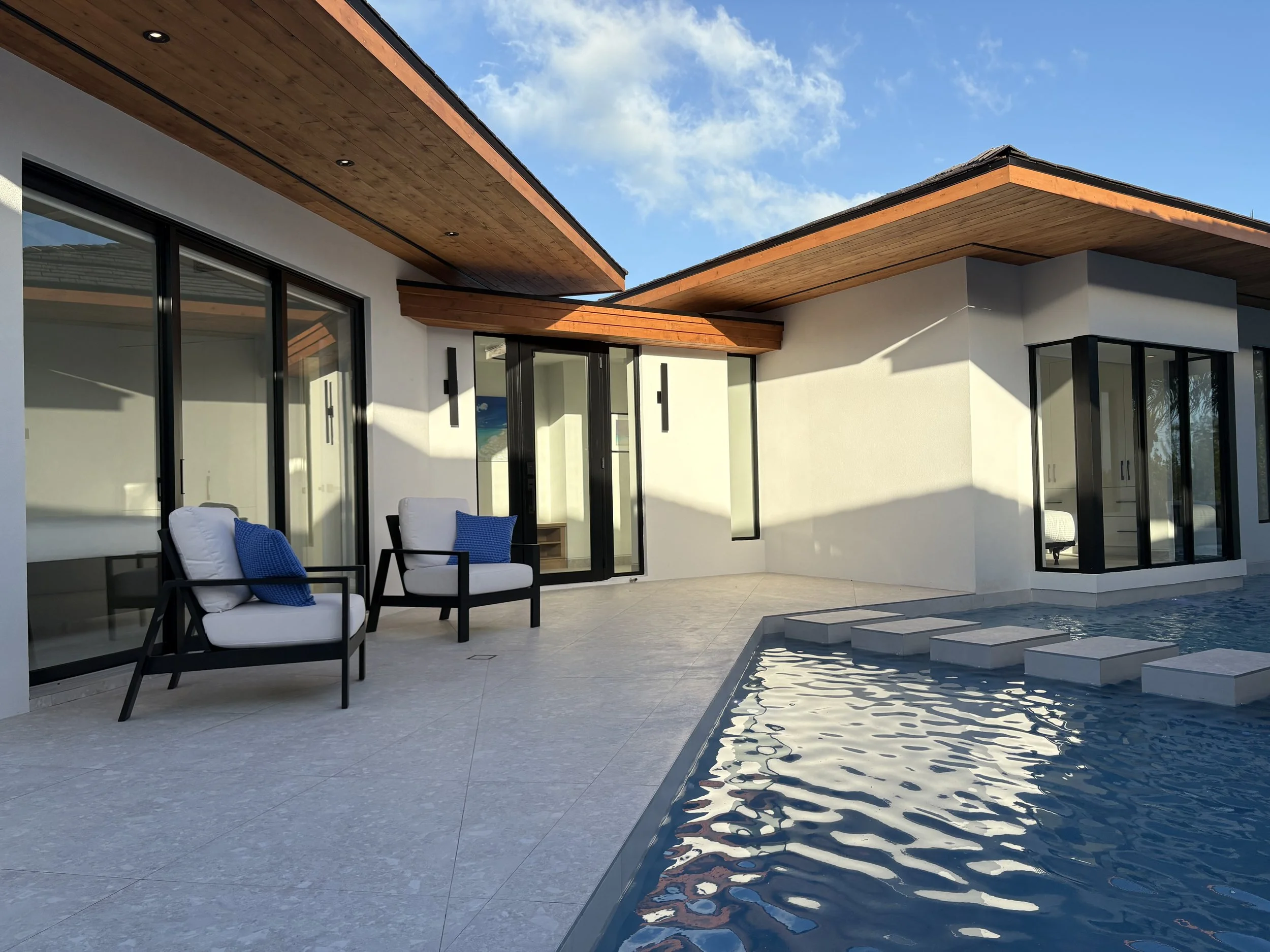 Modern house with large glass sliding doors, a pool with stepping stones, and outdoor seating with blue pillows under a wooden overhang.