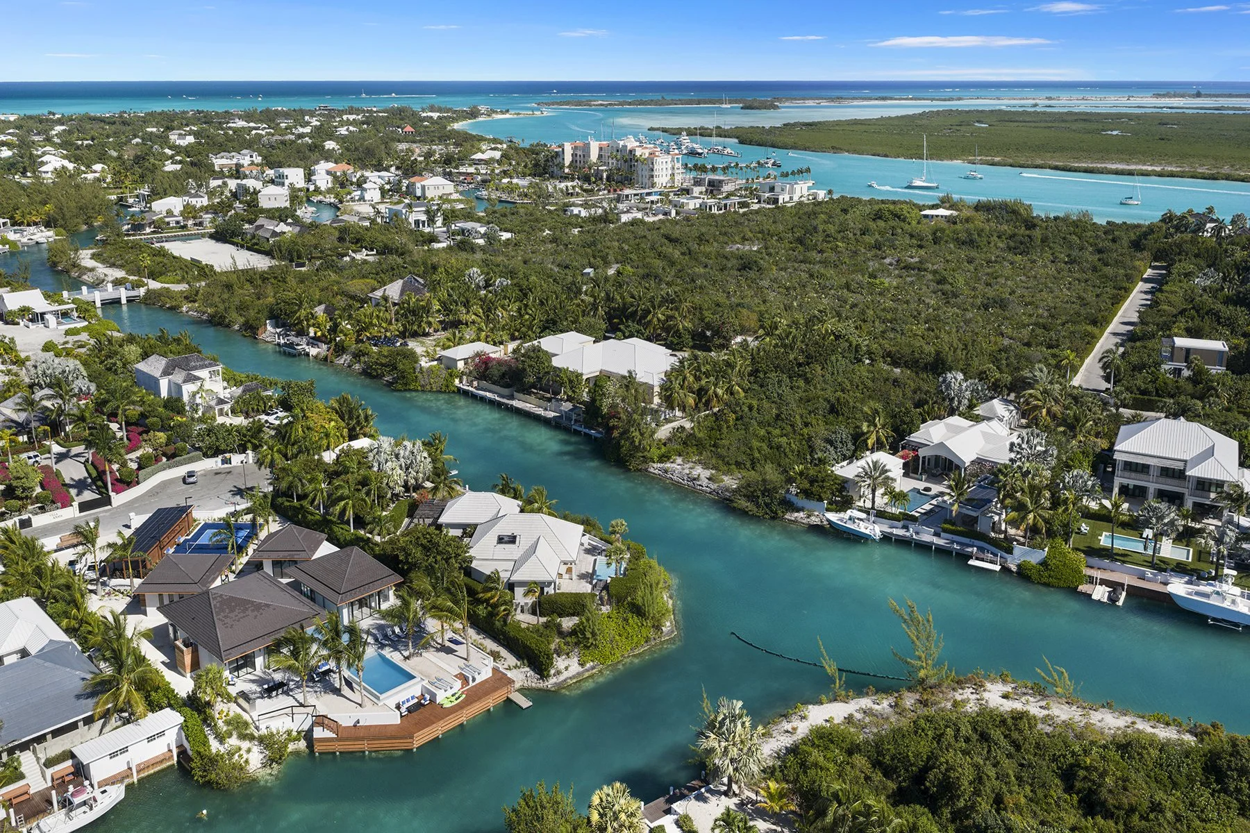 Aerial view of a tropical waterfront community with houses, lush greenery, canals, boats, and a distant ocean.