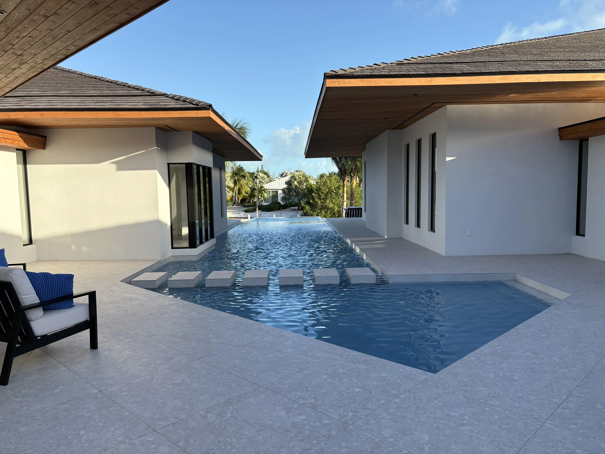 Modern backyard with a private swimming pool, gray tile flooring, white buildings with wooden accents, outdoor seating with blue cushions, and a clear blue sky.