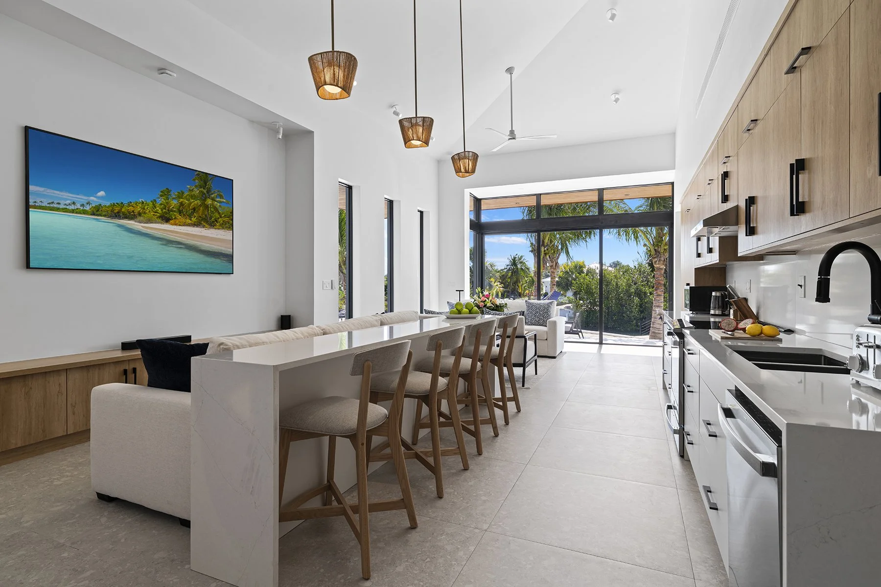 Modern open-concept kitchen and living area with white walls, wooden cabinetry, a large sliding glass door leading to an outdoor patio, and tropical trees outside.