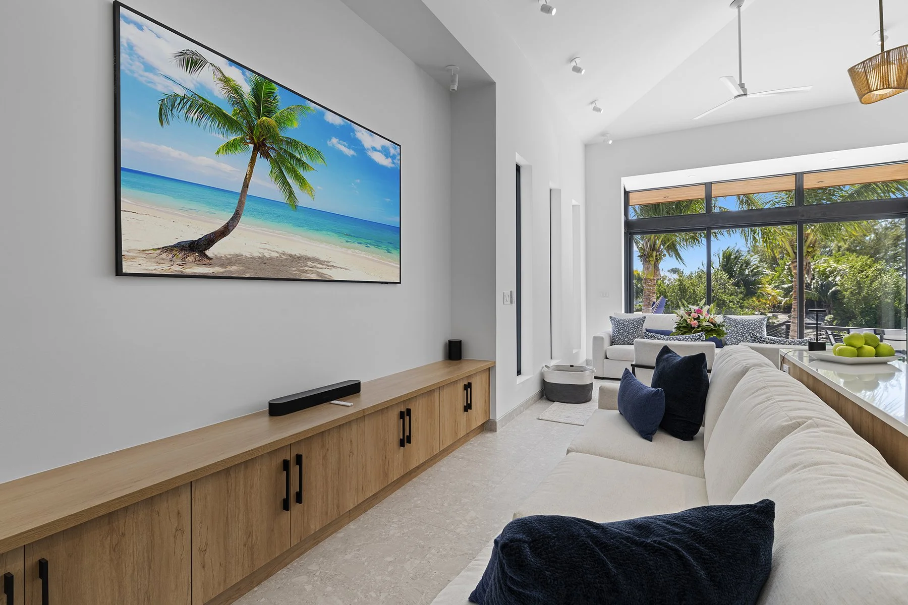 Living room with a large wall-mounted TV displaying a beach scene, white sofa with dark pillows, and large windows overlooking a tropical outdoor area.