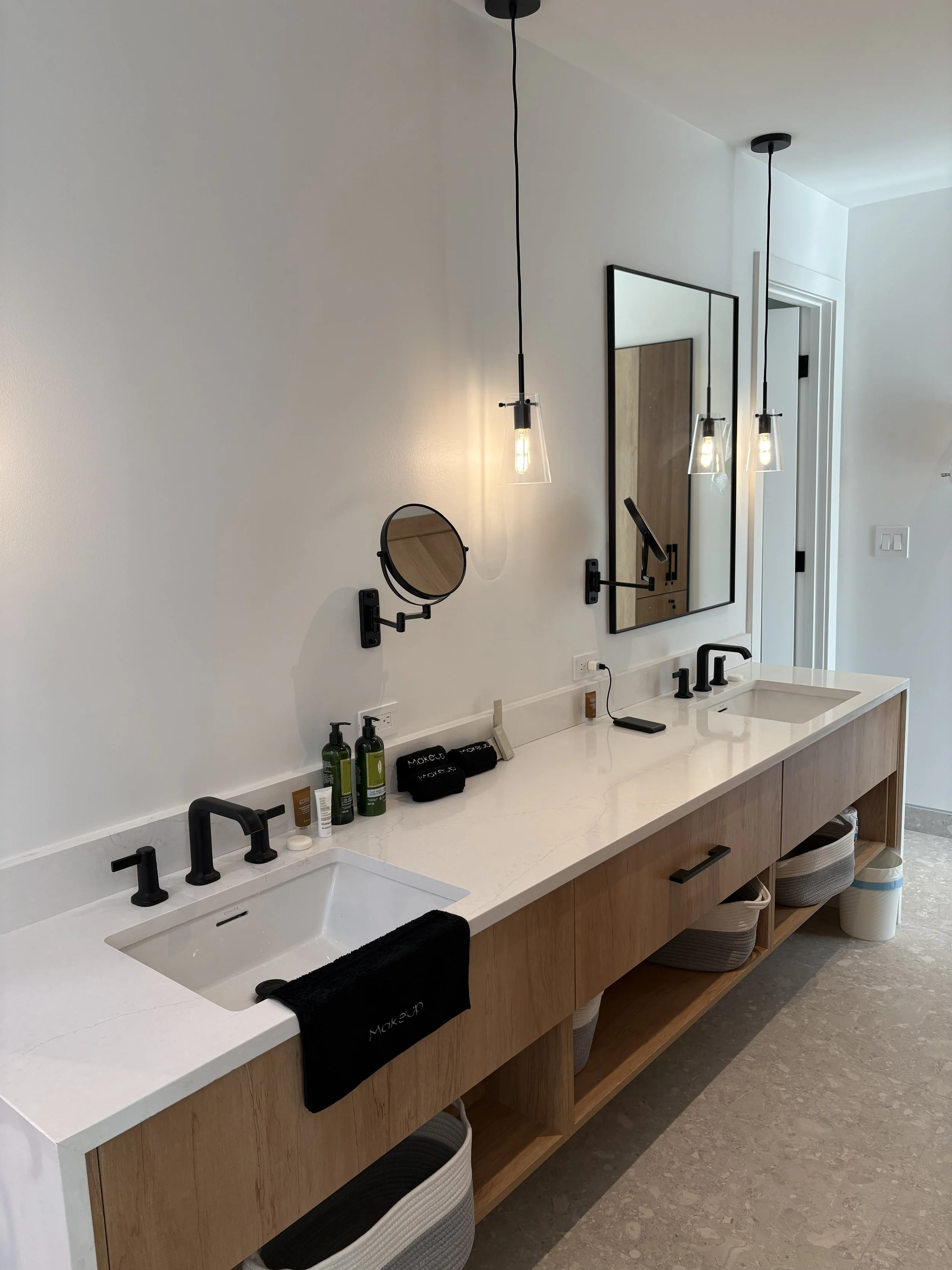 Modern bathroom vanity with a double sink, black fixtures, and a wooden cabinet, decorated with toiletries and towels, illuminated by hanging pendant lights.