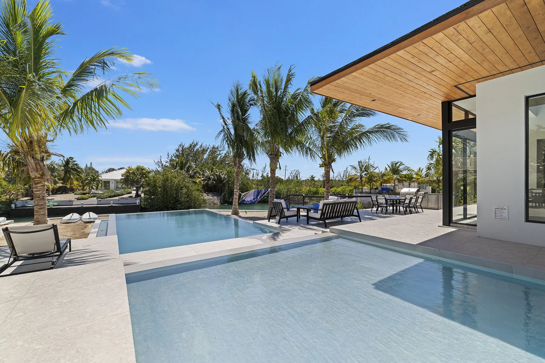 Modern backyard with a swimming pool, lounge chairs, palm trees, and outdoor seating under a wooden overhang.