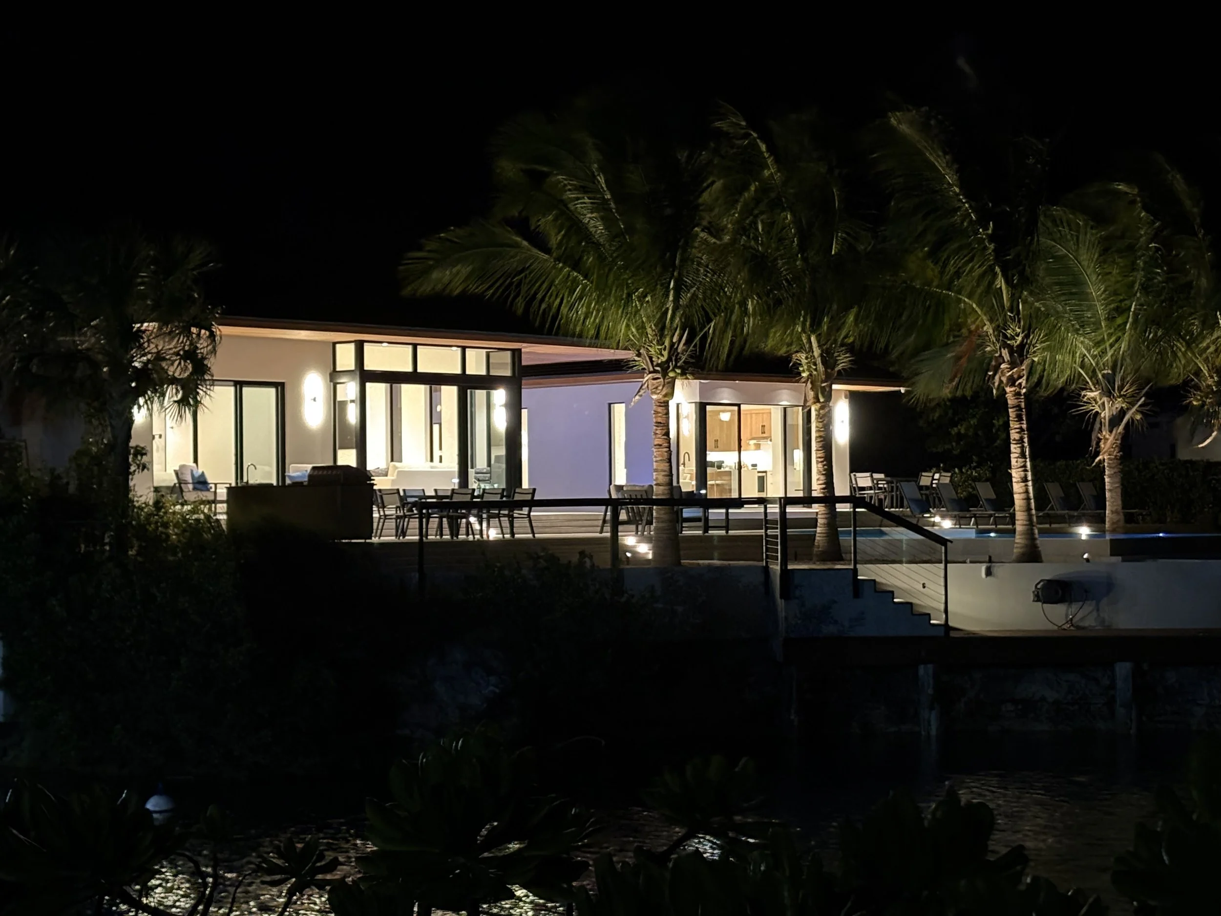 Nighttime view of a modern house with large windows, surrounded by palm trees, illuminated outdoor lighting, and a deck with tables and chairs.