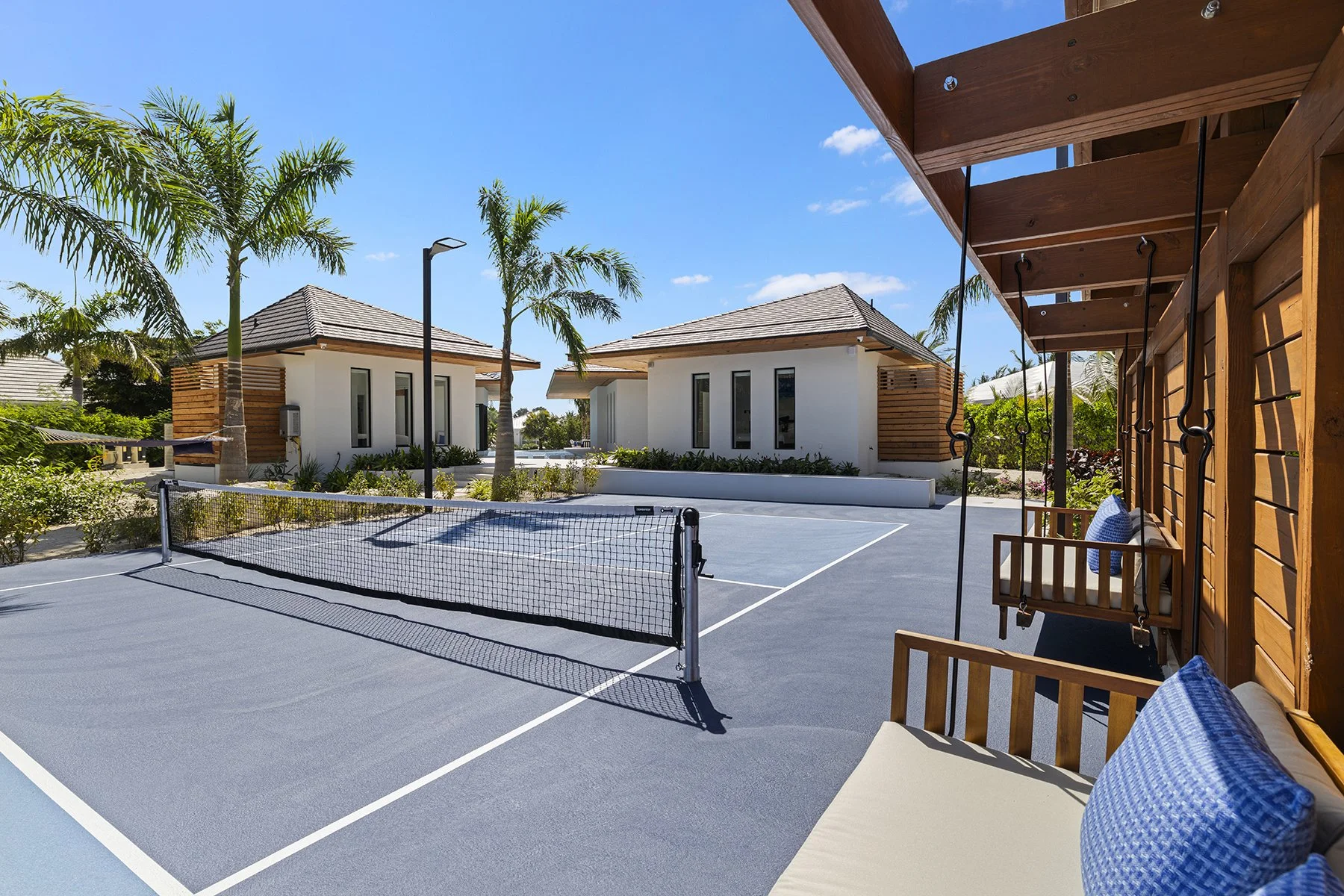 Tennis court with a net, surrounded by palm trees, modern white buildings with wooden accents, sunny sky, and a swing with blue pillows on a wooden structure.