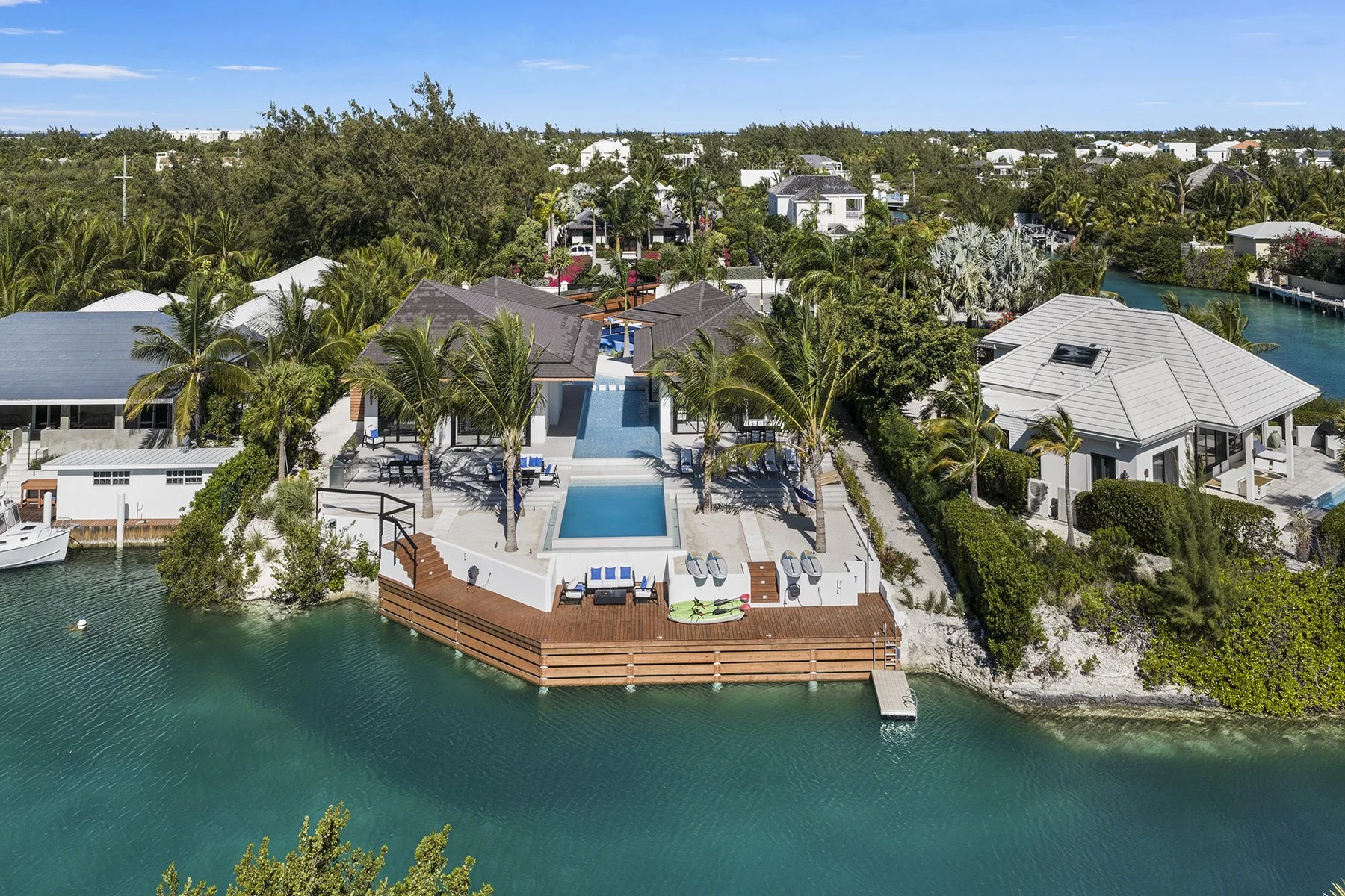 Aerial view of a waterfront modern house with swimming pool, surrounded by palm trees, with boats docked nearby in a lush tropical neighborhood.