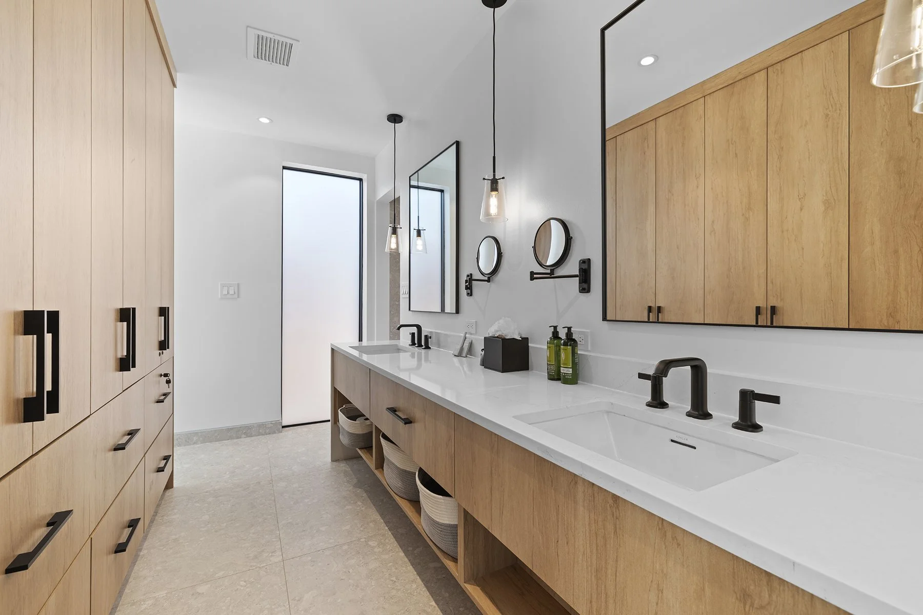 Modern bathroom with wooden cabinets, white countertop, black faucets, large mirror, wall-mounted magnifying mirrors, pendant lights, and beige tiled floor.