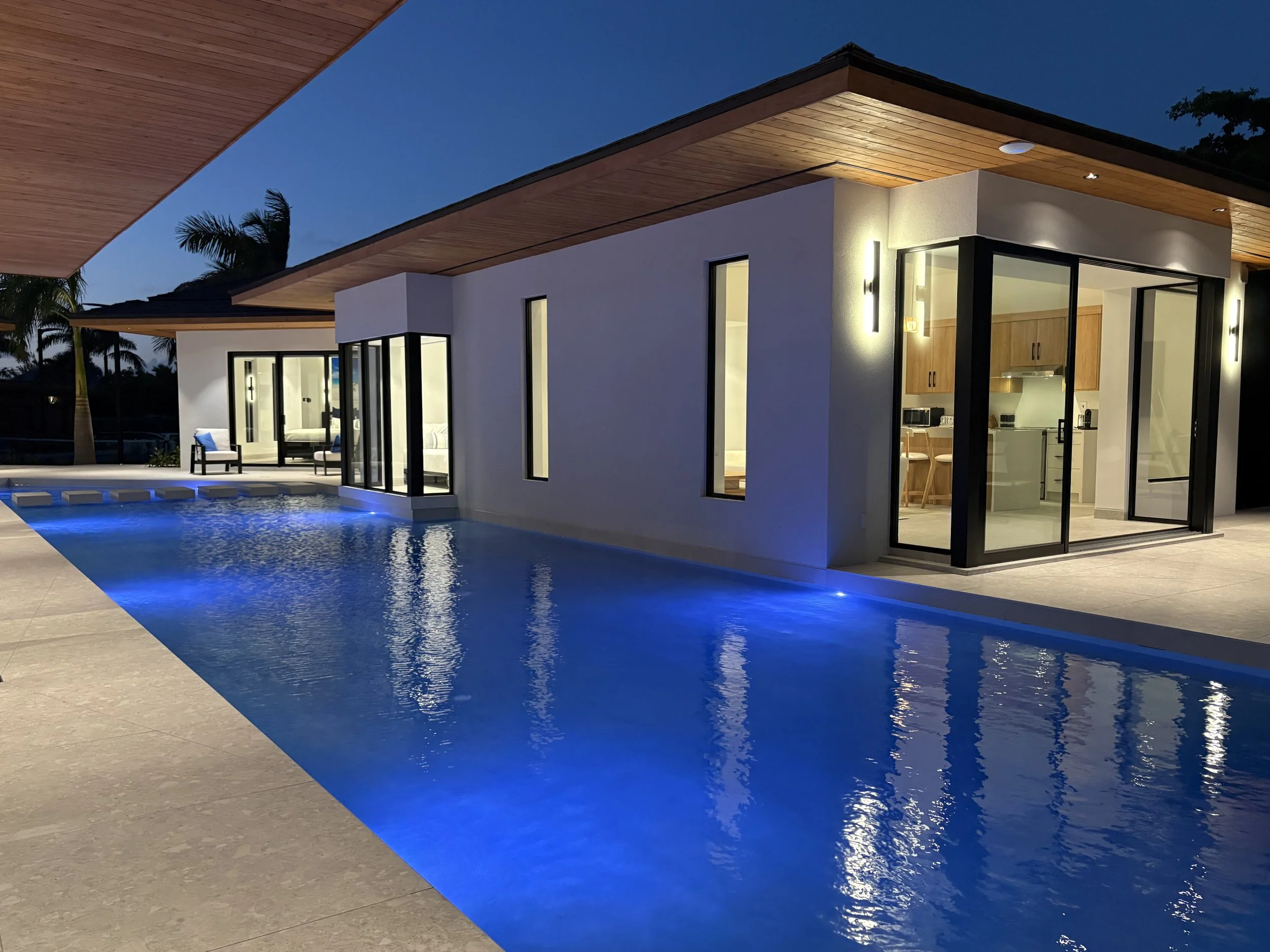 Modern house with large windows and an outdoor pool at dusk, illuminated by exterior lights, with a visible interior kitchen and living space.