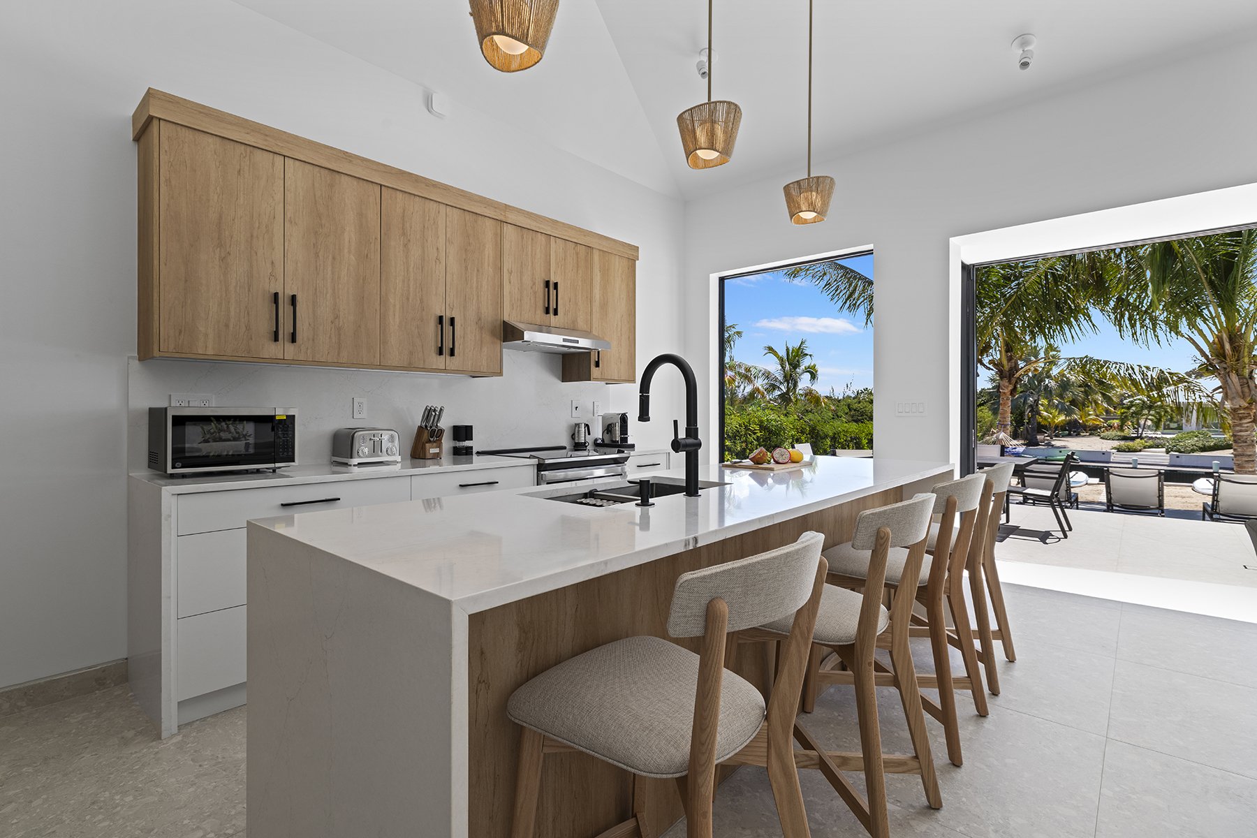 Modern kitchen with white walls, wooden cabinets, white countertop, black faucet, appliances, and a view of outdoor patio with trees and blue sky.
