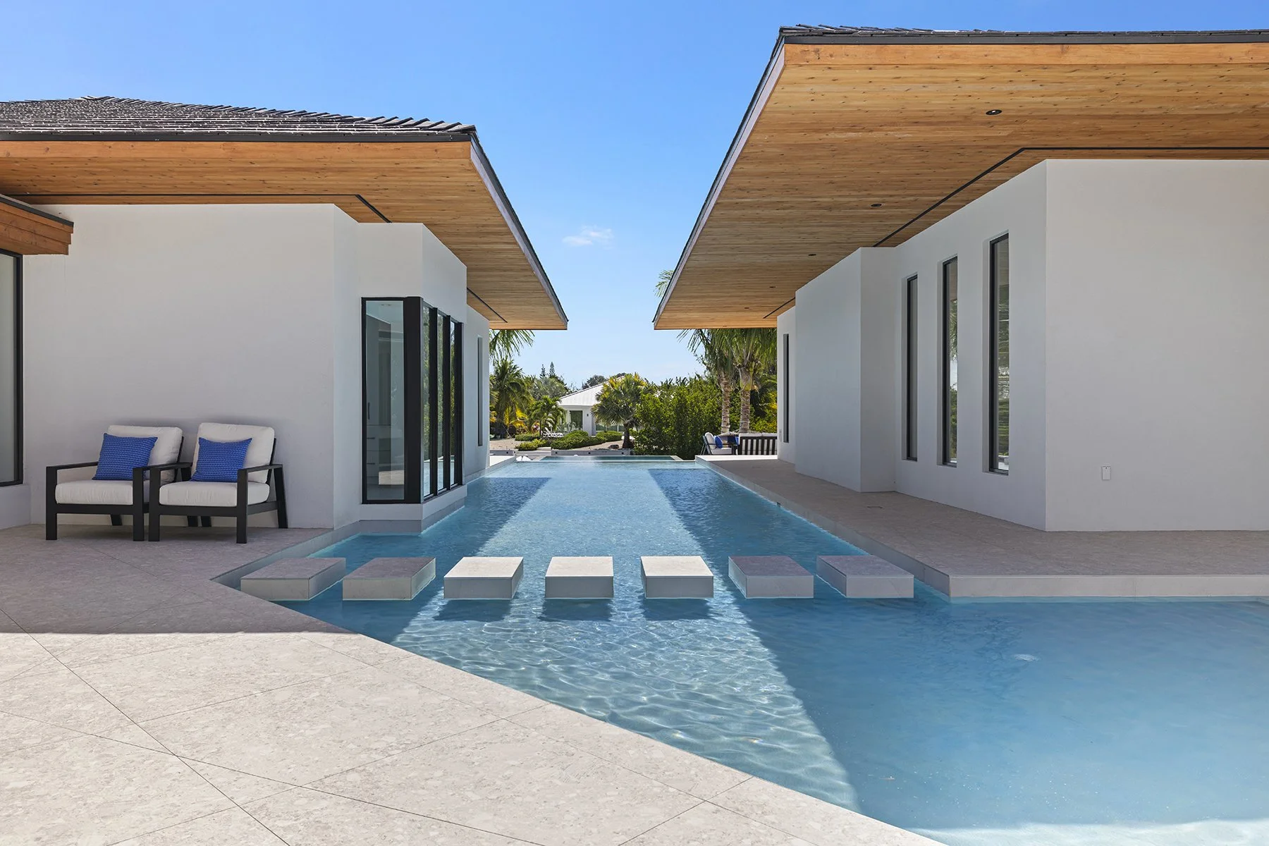 Modern house with a swimming pool featuring floating stepping stones, white walls, wooden roof accents, and outdoor seating with blue cushions, surrounded by a landscaped yard with palm trees.
