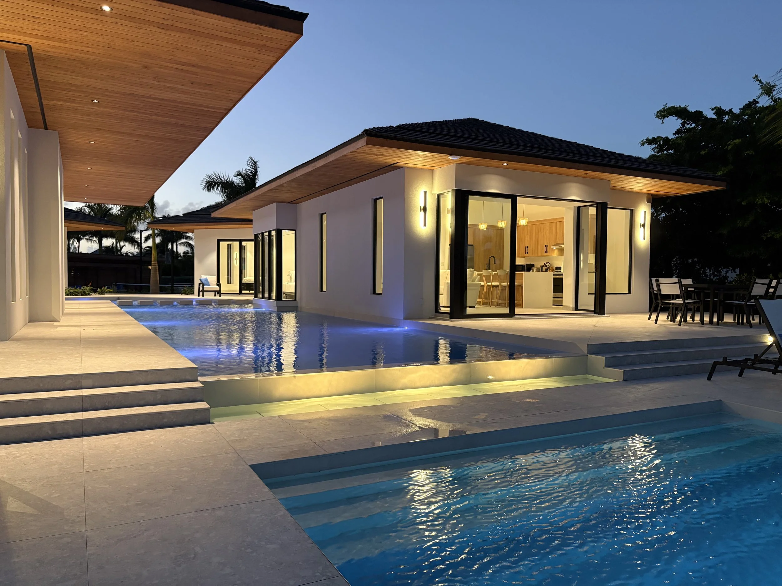 Modern house with a swimming pool and outdoor seating area at dusk, large glass windows, wood accents, and tropical trees in the background.