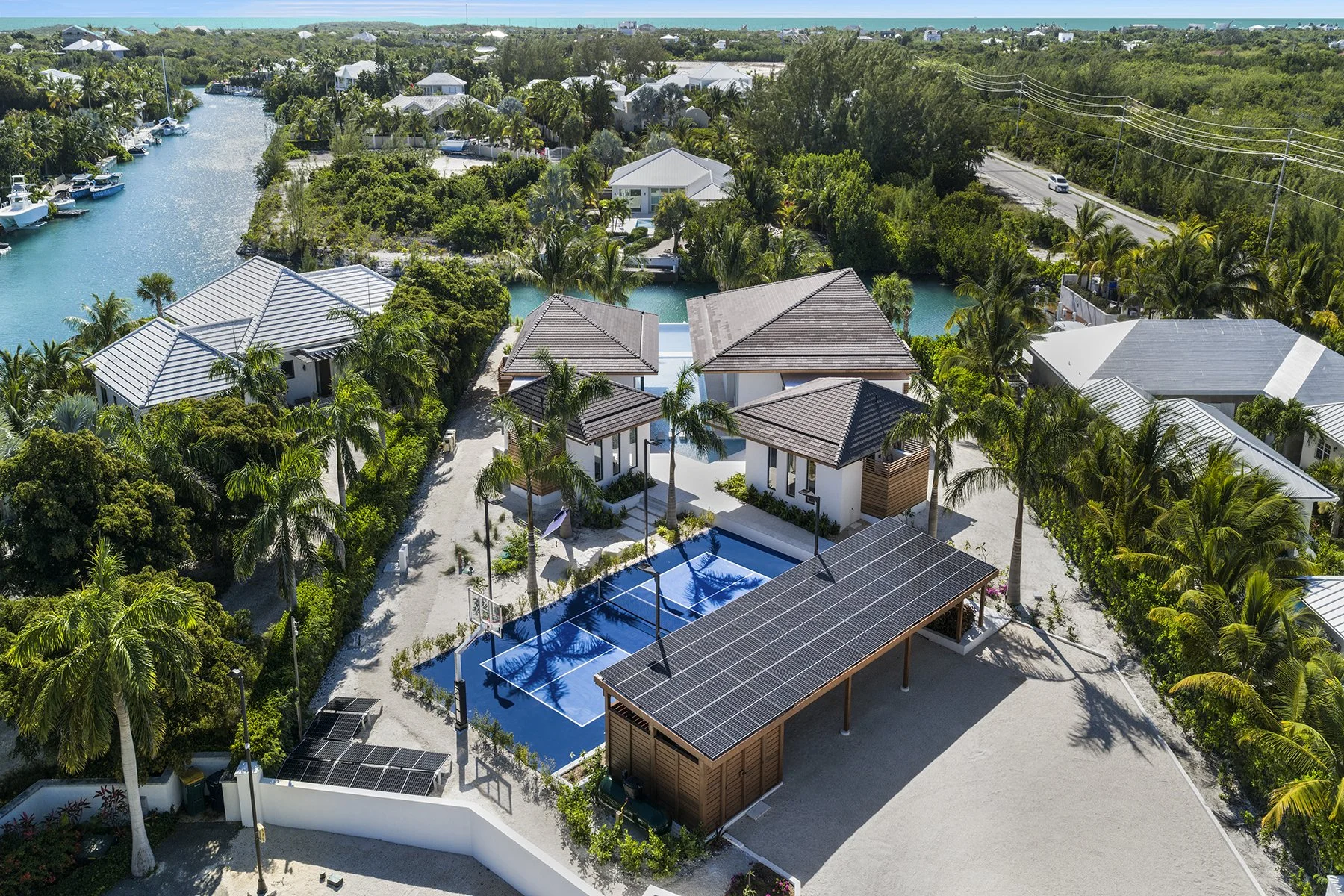 Aerial view of a tropical residential neighborhood featuring a house with a solar-paneled carport, a swimming pool, palm trees, and nearby water channels with boats and houses along the water, surrounded by lush greenery and ocean in the background.
