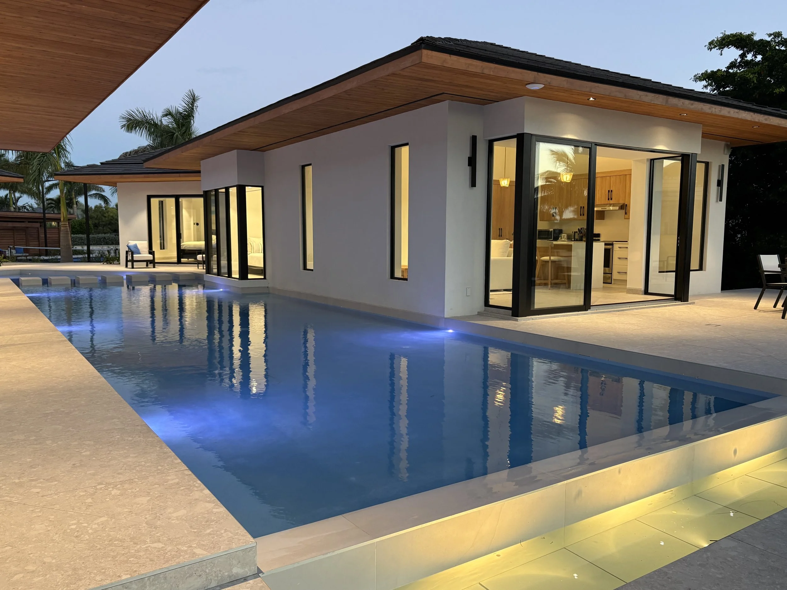 Modern house with large glass sliding doors, a swimming pool with blue LED lighting, and outdoor seating. The house has a flat roof with wooden accents and is illuminated from inside.