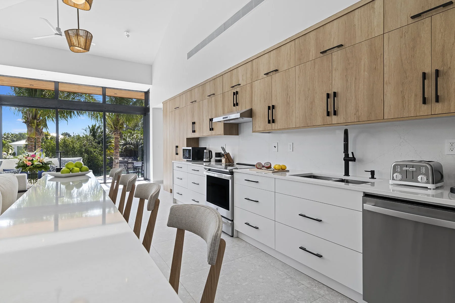 Modern kitchen with white and wood cabinets, black handles, and a large window with palm trees outside.