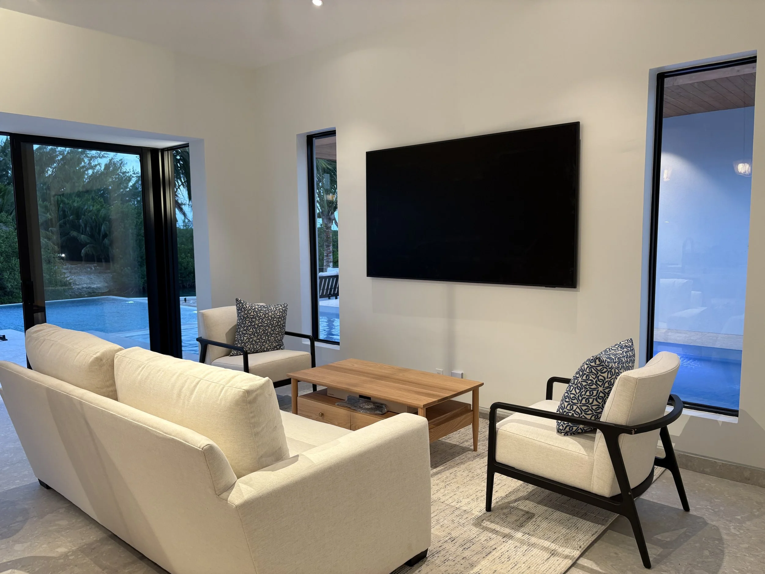 Living room with a beige sofa, two armchairs with patterned pillows, a wooden coffee table, large windows showing outdoor pool and trees, and a wall-mounted TV.