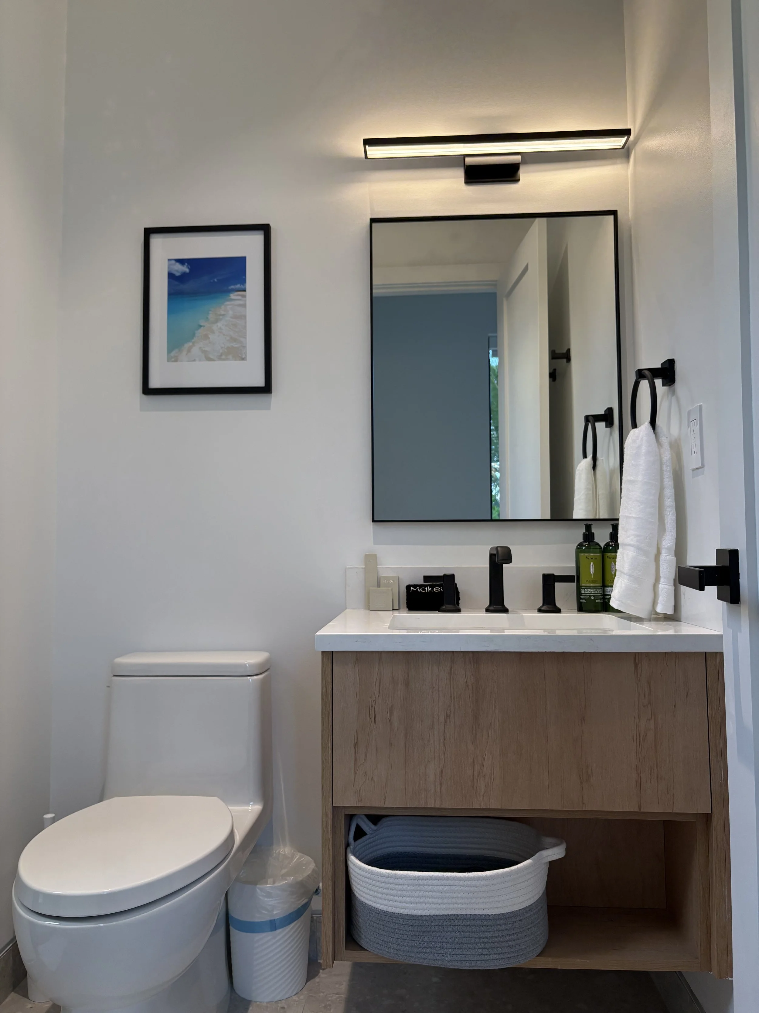 Modern bathroom with a white toilet, a wooden vanity with a white countertop, a large mirror, framed beach photo, and black fixtures.