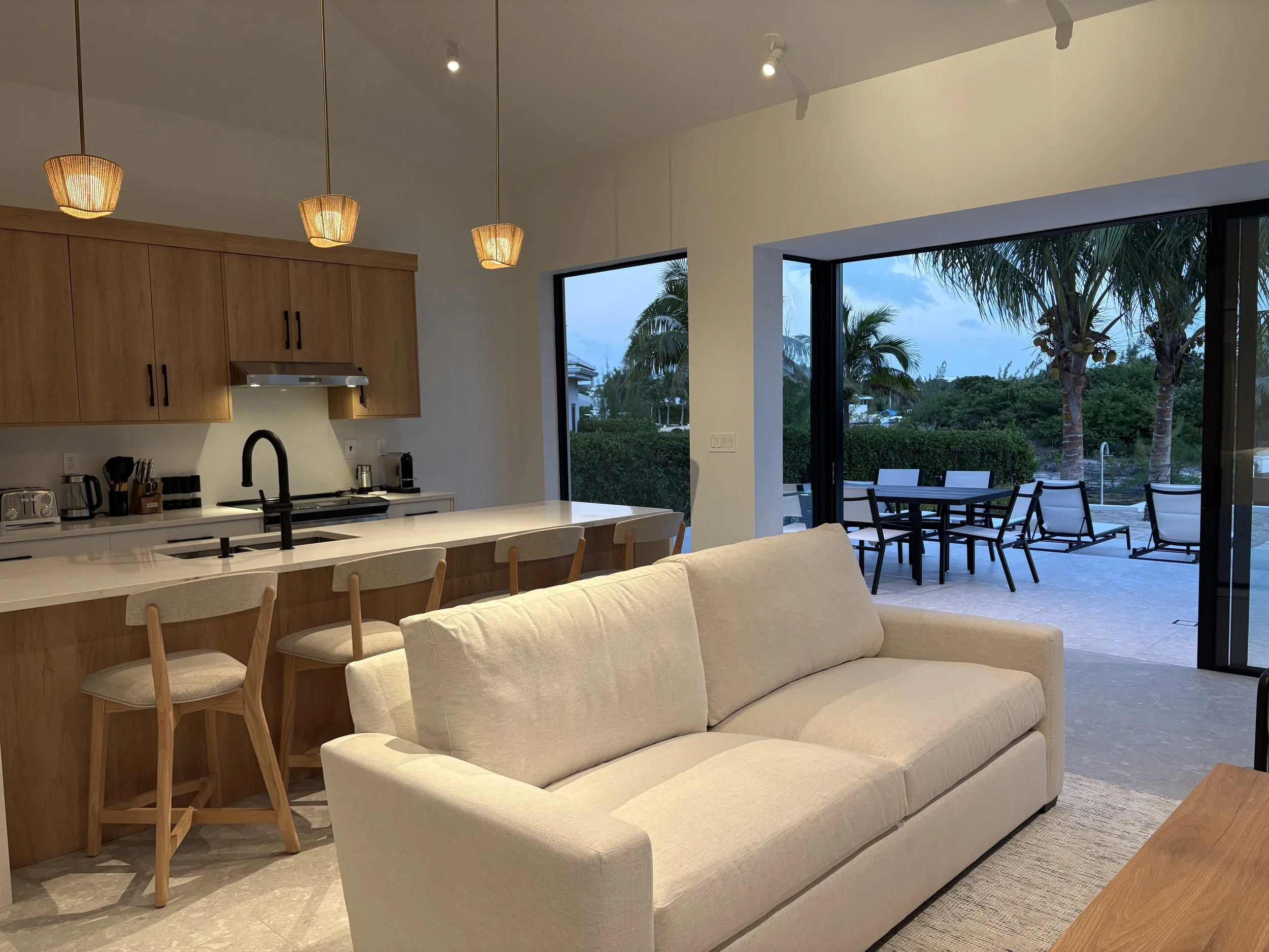 Modern living room with beige sofa, wooden kitchen cabinets, white kitchen island with bar stools, sliding glass door opening to outdoor patio with tables and chairs, palm trees in the background.