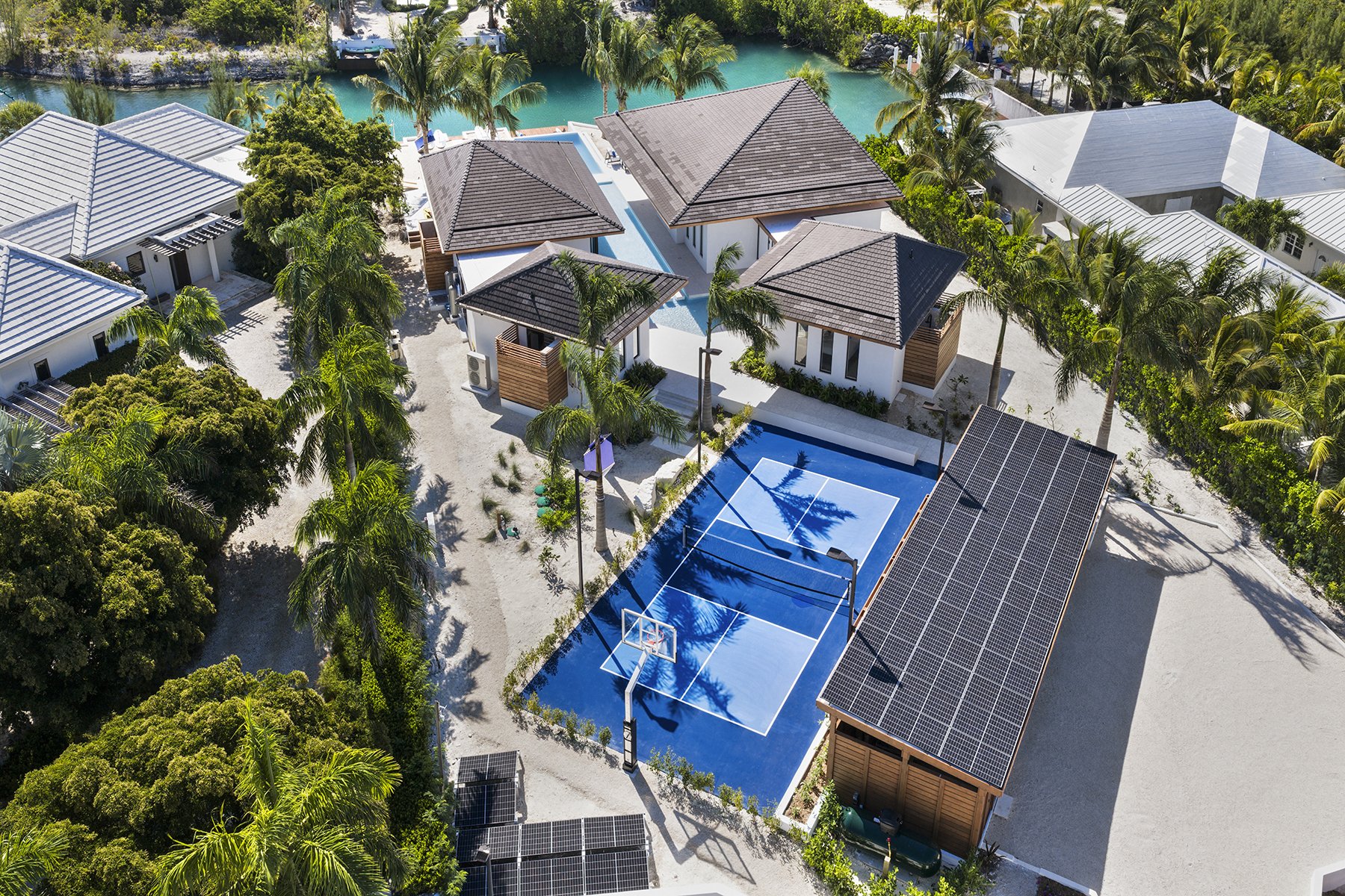 Aerial view of a tropical resort with white buildings, palm trees, a swimming pool, a tennis court, and solar panels on a large building roof, surrounded by greenery.