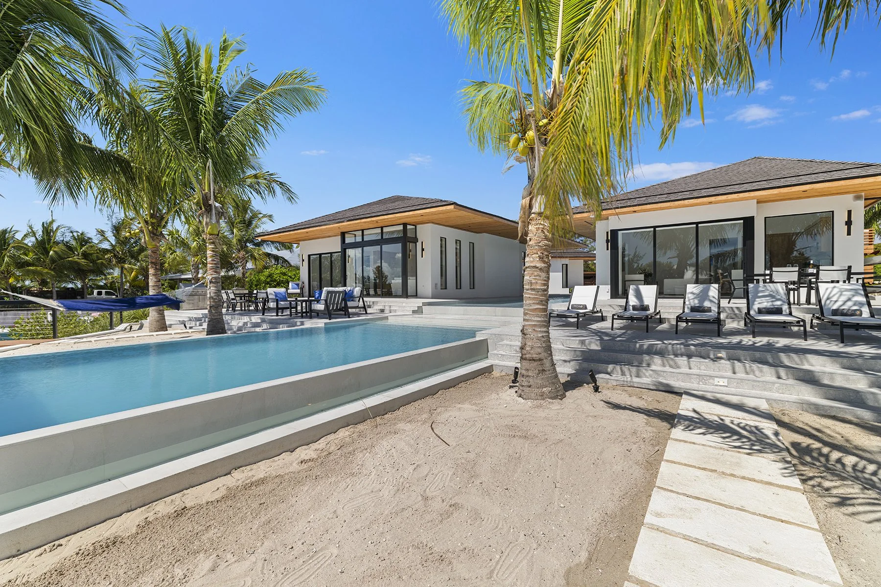 Luxury modern house with a swimming pool in a tropical setting, featuring palm trees, outdoor seating, and a clear blue sky.