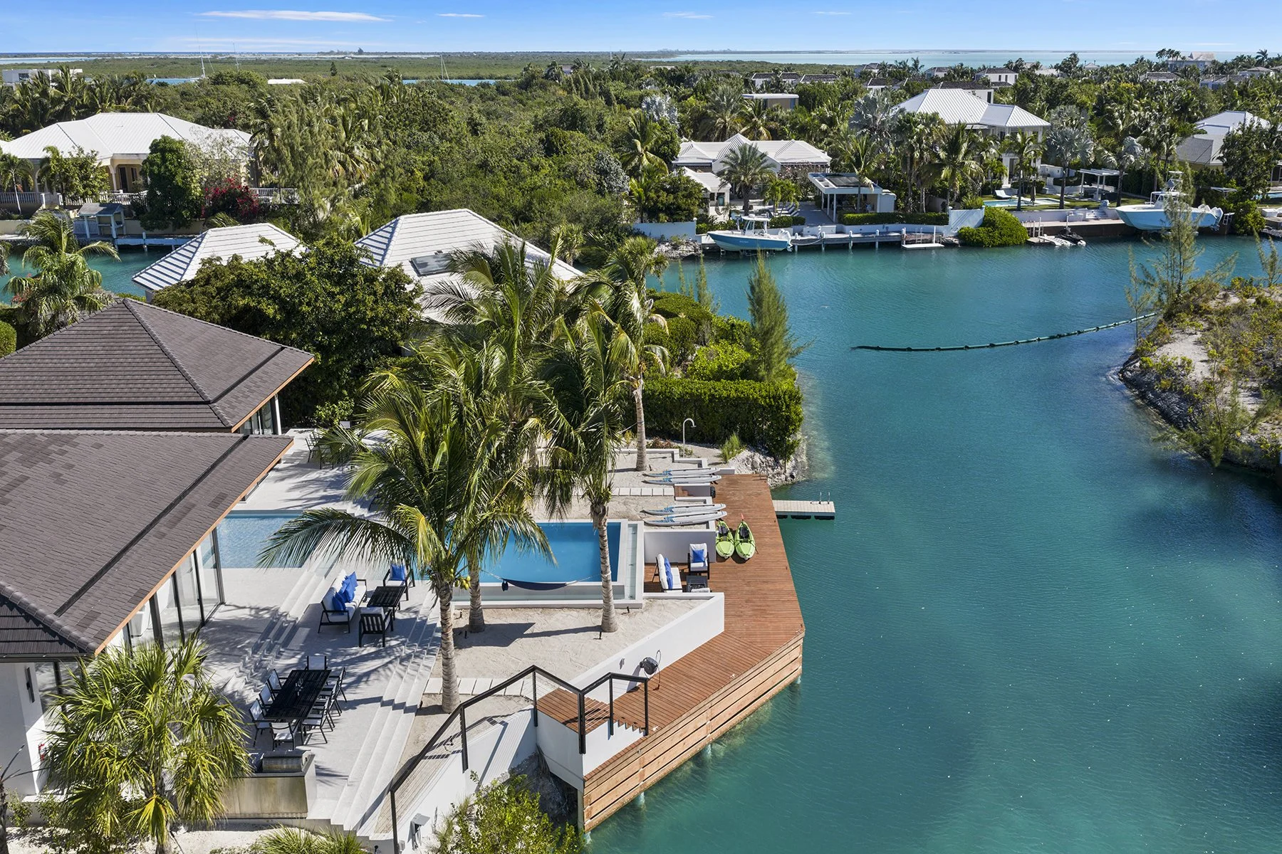 Aerial view of waterfront homes with pools, palm trees, and boats on a clear turquoise waterway in a sunny tropical setting.