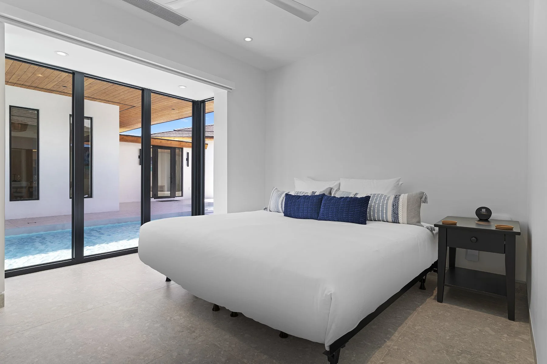 Modern bedroom with white bed, decorative pillows, black nightstand, and floor-to-ceiling glass doors leading to a pool and outdoor patio.