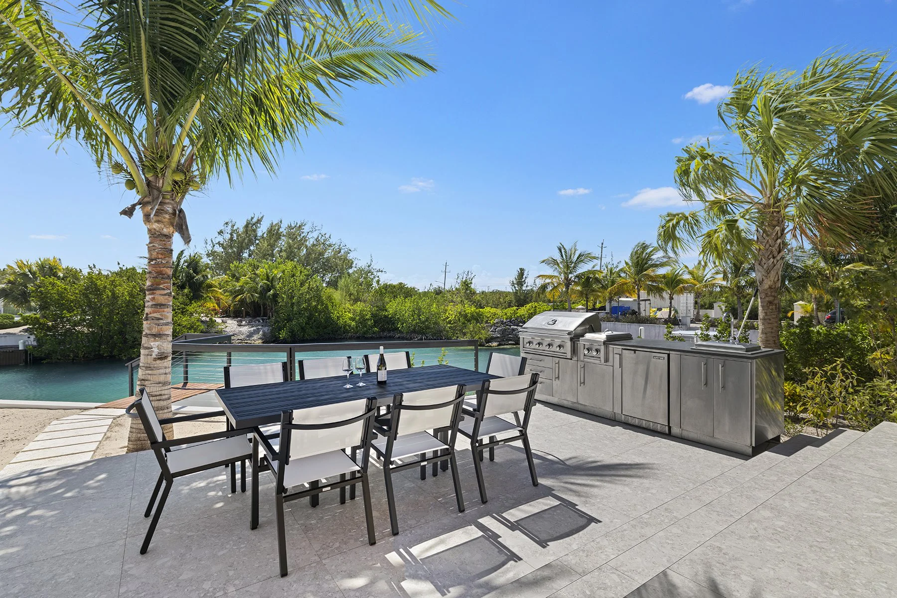 Outdoor patio with a dining table, chairs, and a built-in grill, overlooking a tropical waterway with palm trees and lush greenery under a clear blue sky.
