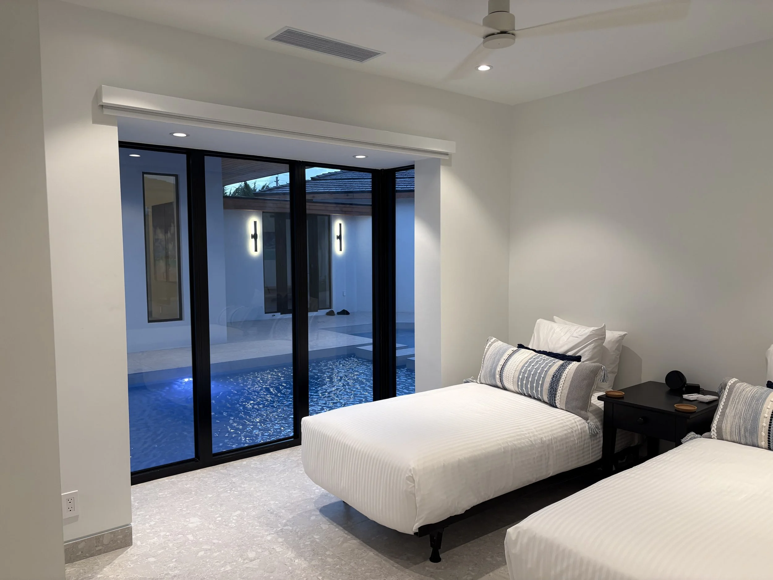 Two twin beds with white bedding and decorative pillows, nightstand with a speaker and small items, sliding glass door leading to a pool area outdoors.